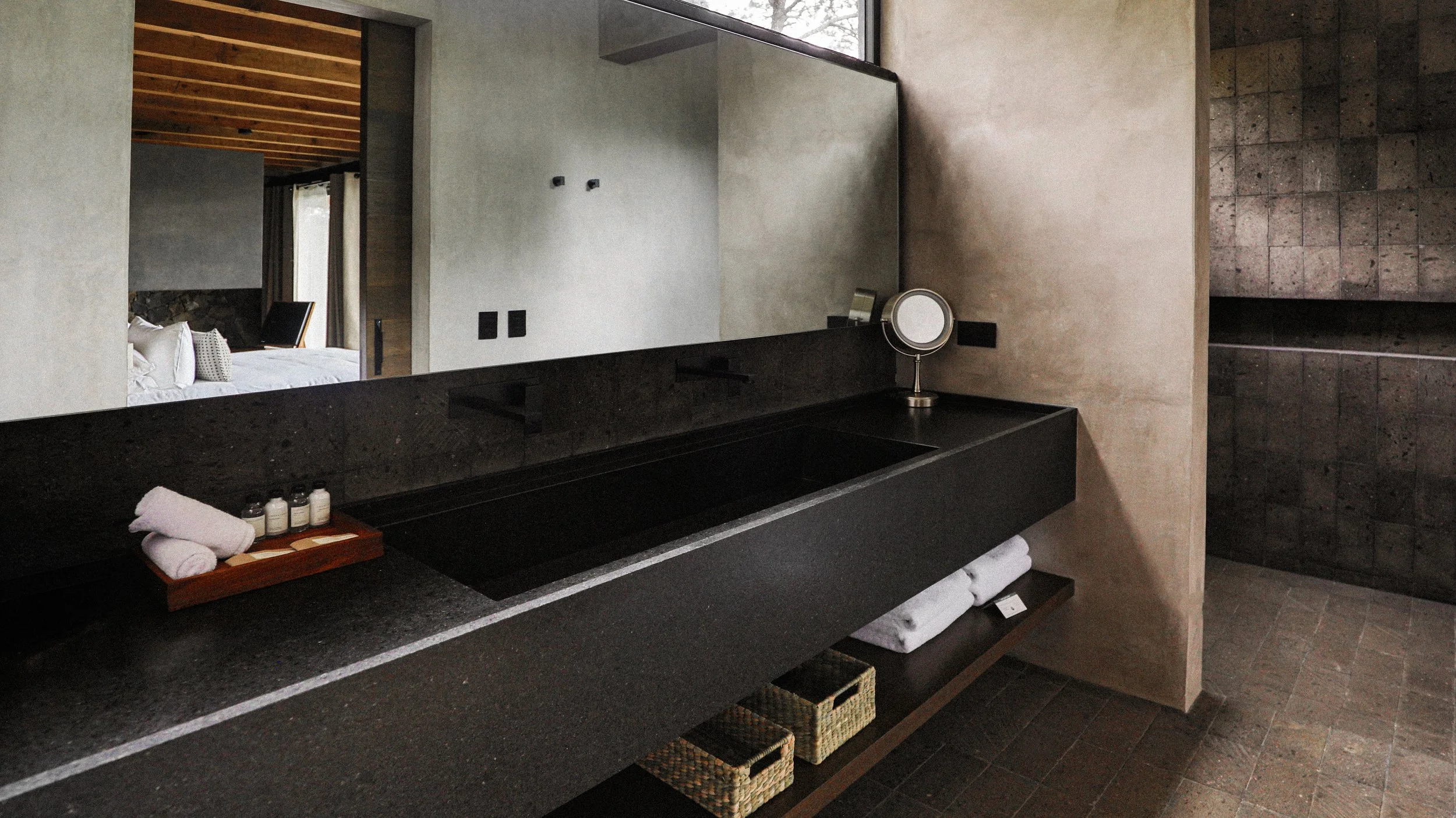 Baño moderno con lavabo de piedra negra, espejo grande y paredes de concreto. Se ve una cama en otra habitación a través de un espejo y hay toallas y pequeños frascos de productos en el lavabo.