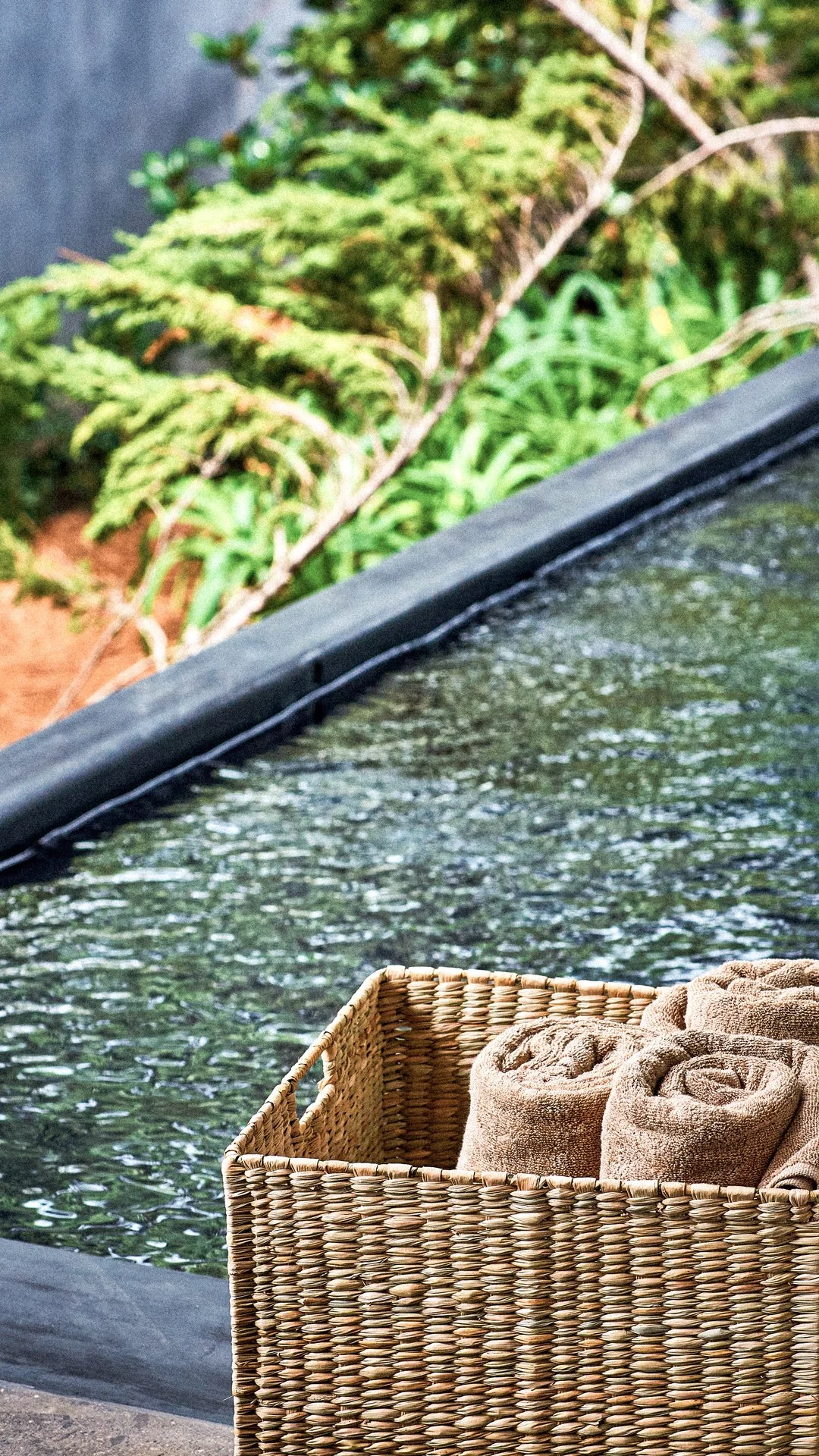Cesta de mimbre con toallas enrolladas junto a una piscina con agua y plantas verdes en el fondo.