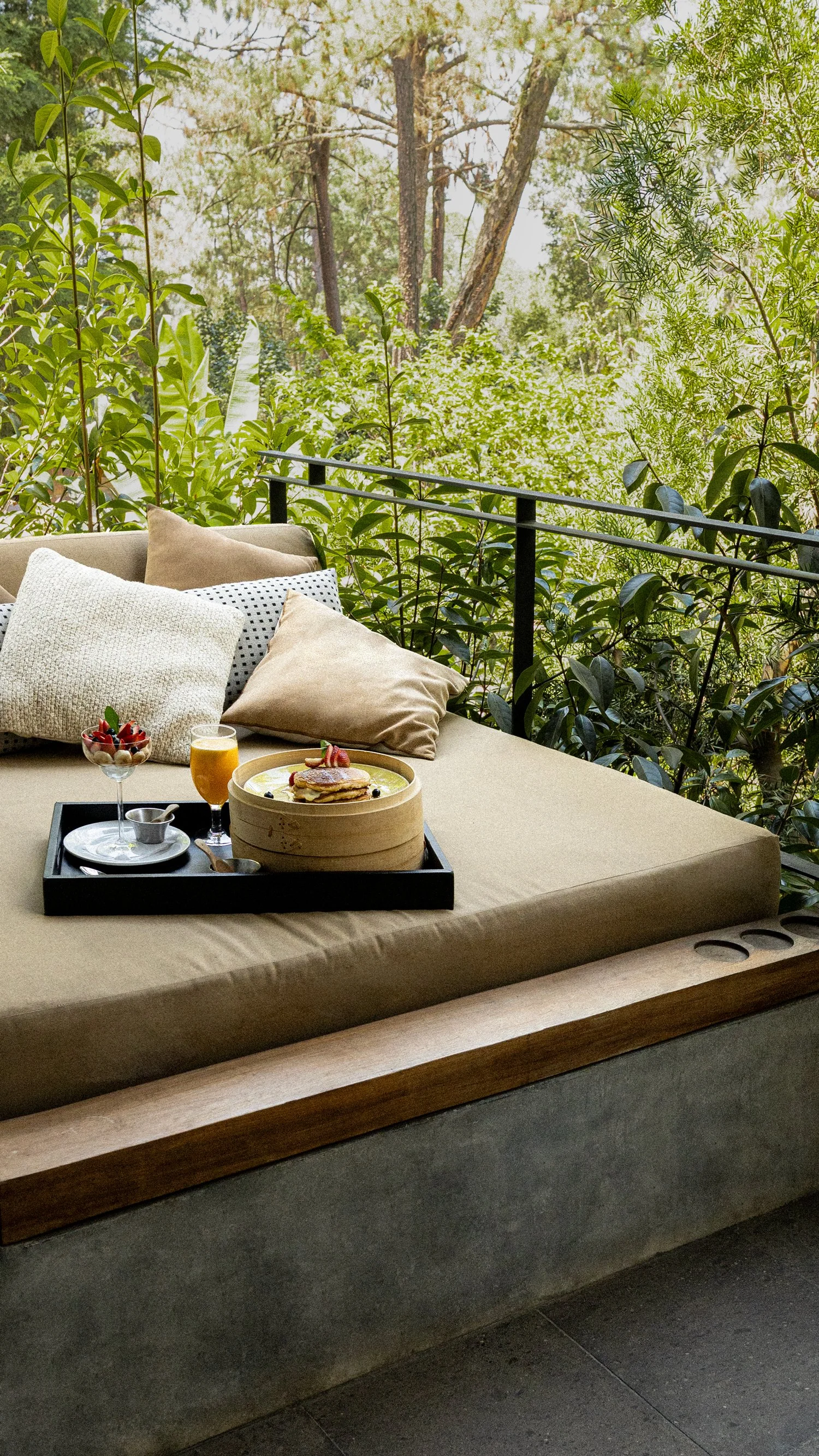 Imagen de un área de descanso con cojines en un sillón junto a un balcón con vista a un bosque de árboles verdes.