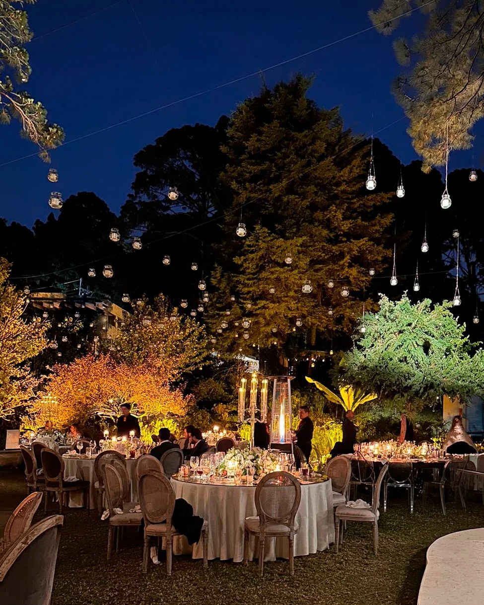 Cena al aire libre con mesas decoradas con velas y flores, luces colgantes y árboles iluminados en un entorno nocturno elegante y festivo.