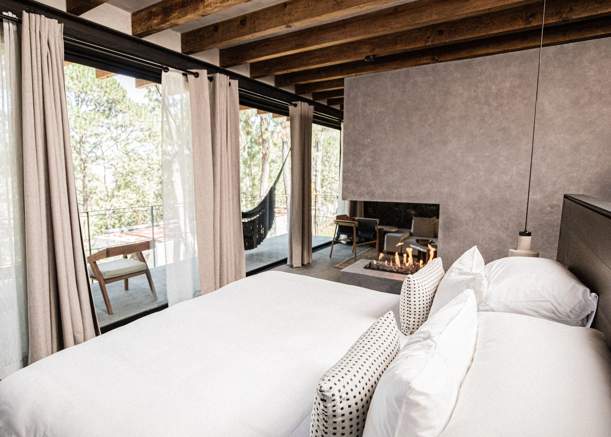 Habitación moderna con cama con sábanas blancas, ventanas grandes con cortinas, balcón con vista a la naturaleza, chimenea encendida y sala con sillones.