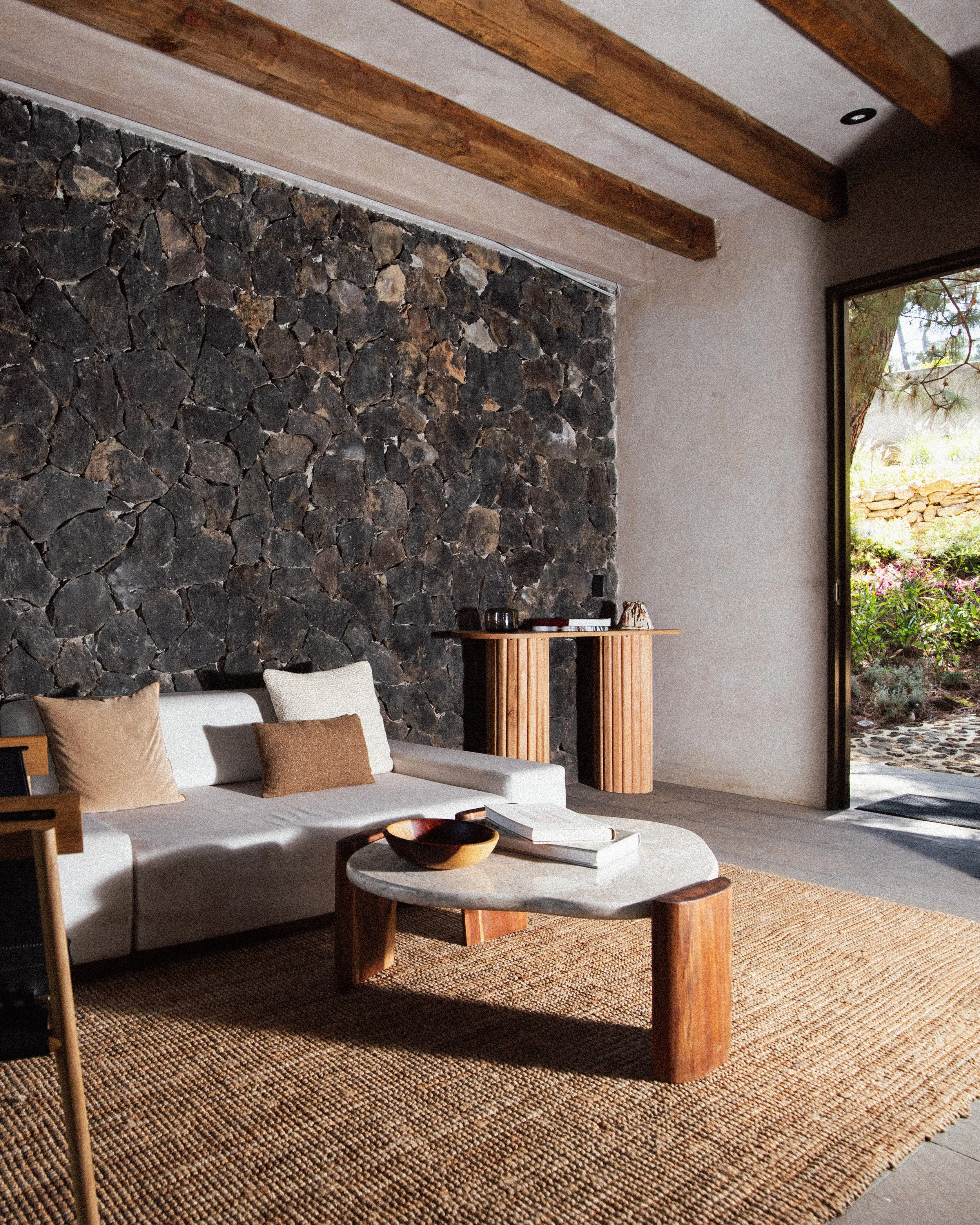 Sala moderna con sofá blanco, cojines beige y marrón, mesa de centro de piedra y madera, pared de piedra negra y vista al exterior con jardín.