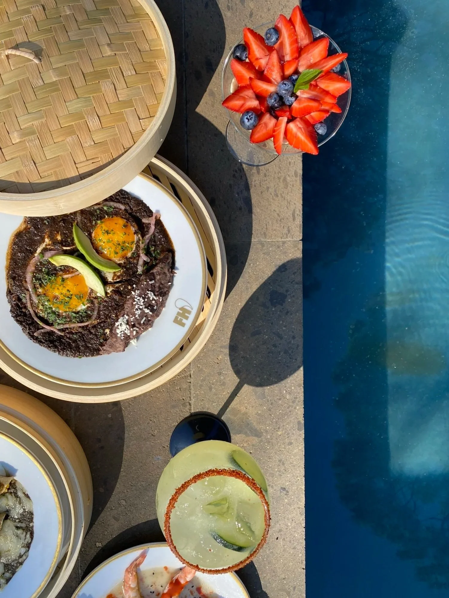 Plato de huevos estrellados con carne de res, acompañado de una bebida con limón, y una copa con fresas y blueberries, todo junto a una piscina.