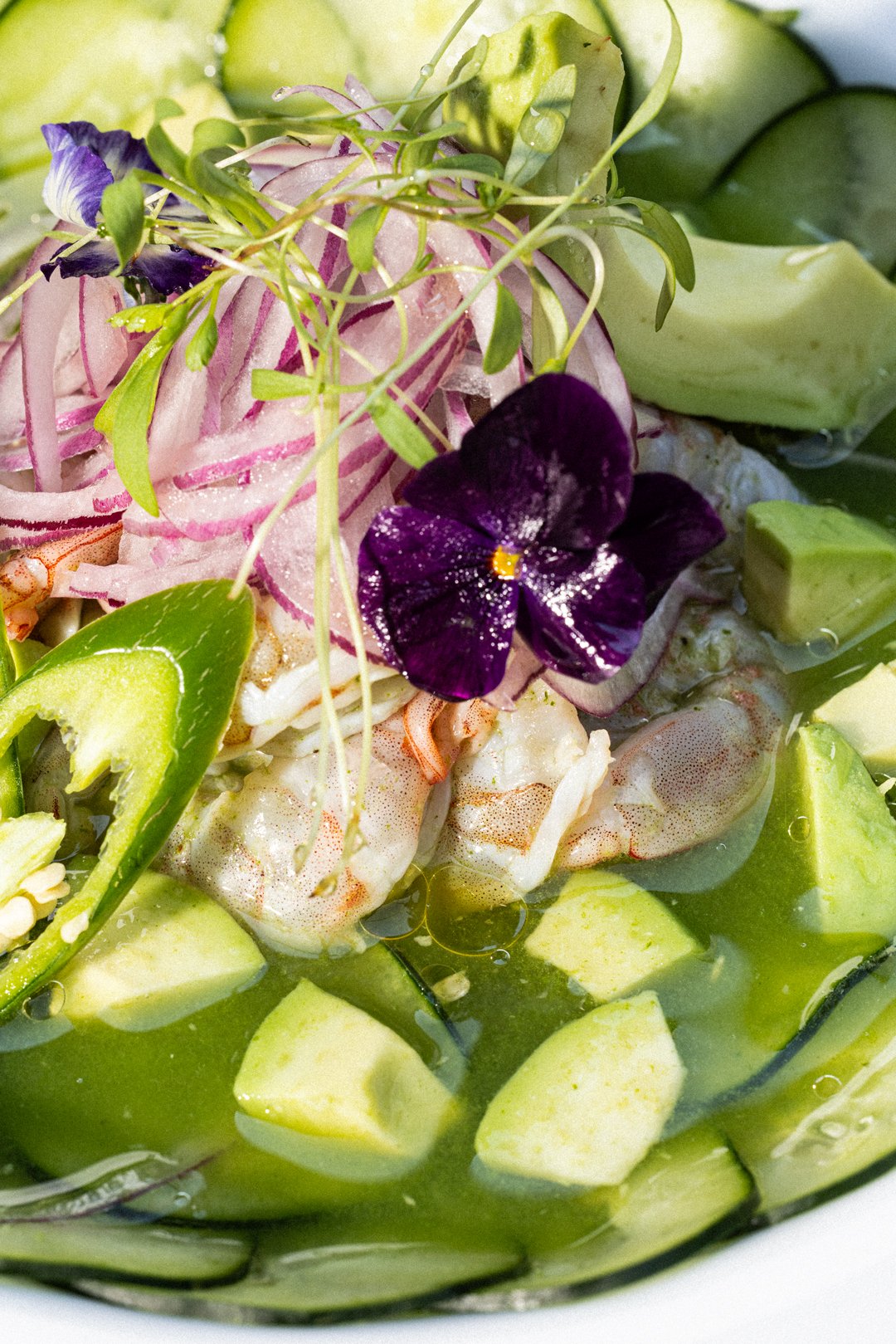 Camarones en una sopa fría con trozos de pepino, cebolla morada, aguacate y decorado con una flor violeta y microverdes.