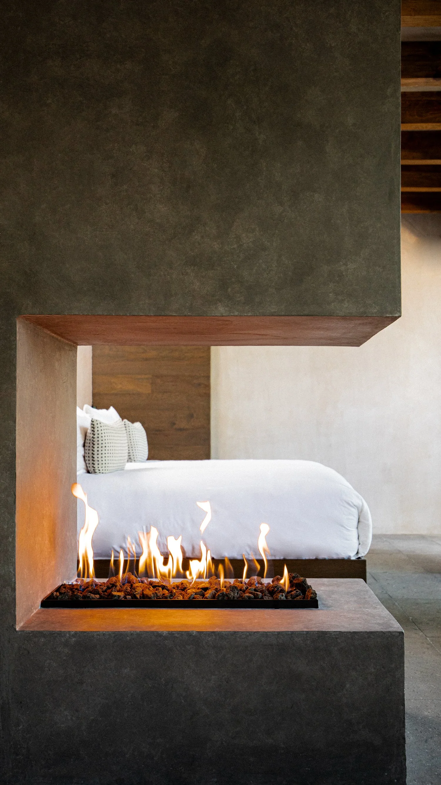 Vista de una cama blanca con cojines, frente a una chimenea moderna de fuego abierto y diseño minimalista en una habitación.