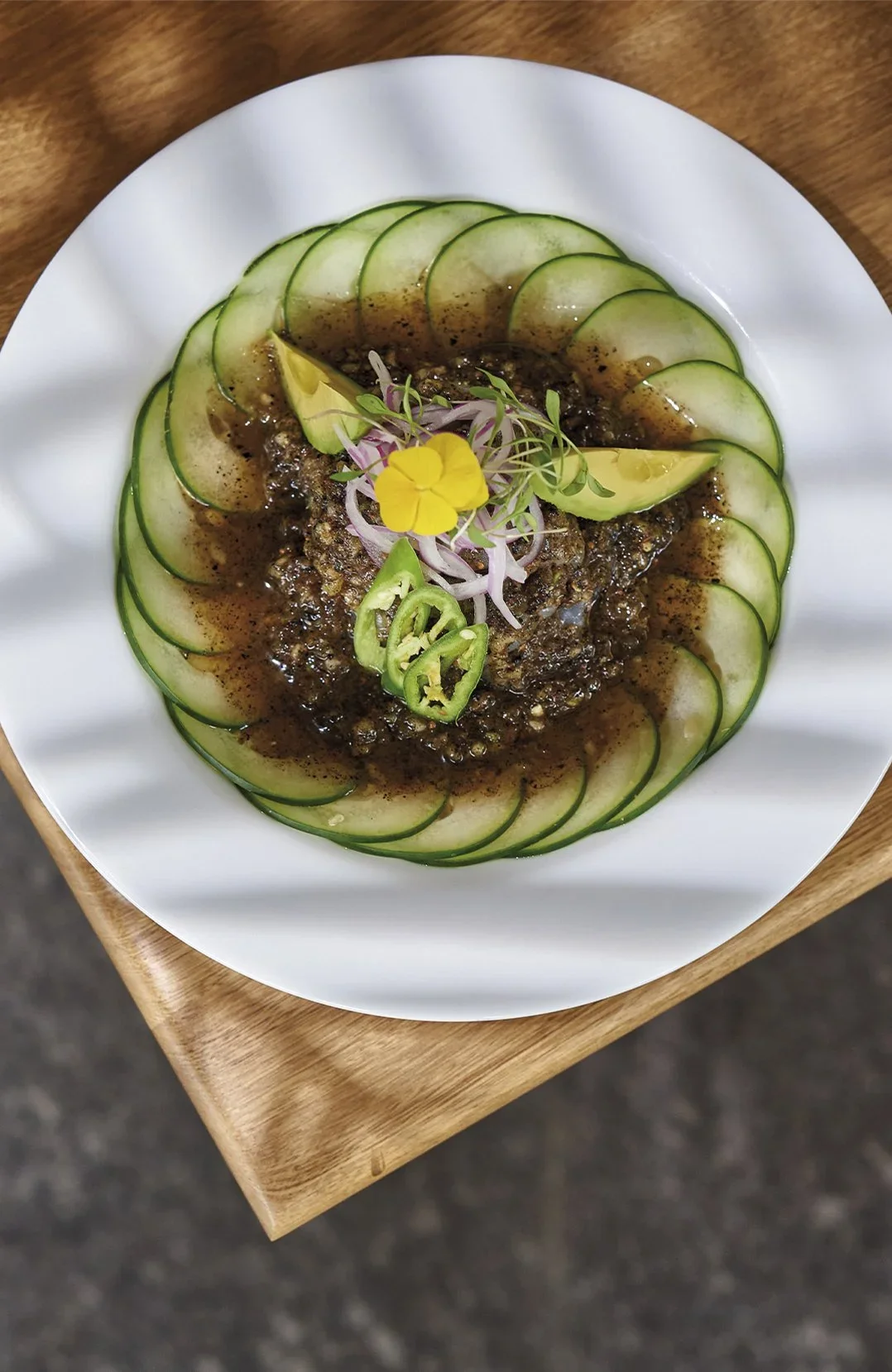 Plato de pescado con salsa oscuro, adornado con rodajas de pepino, aguacate, jalapeños, cebolla morada y flores comestibles.