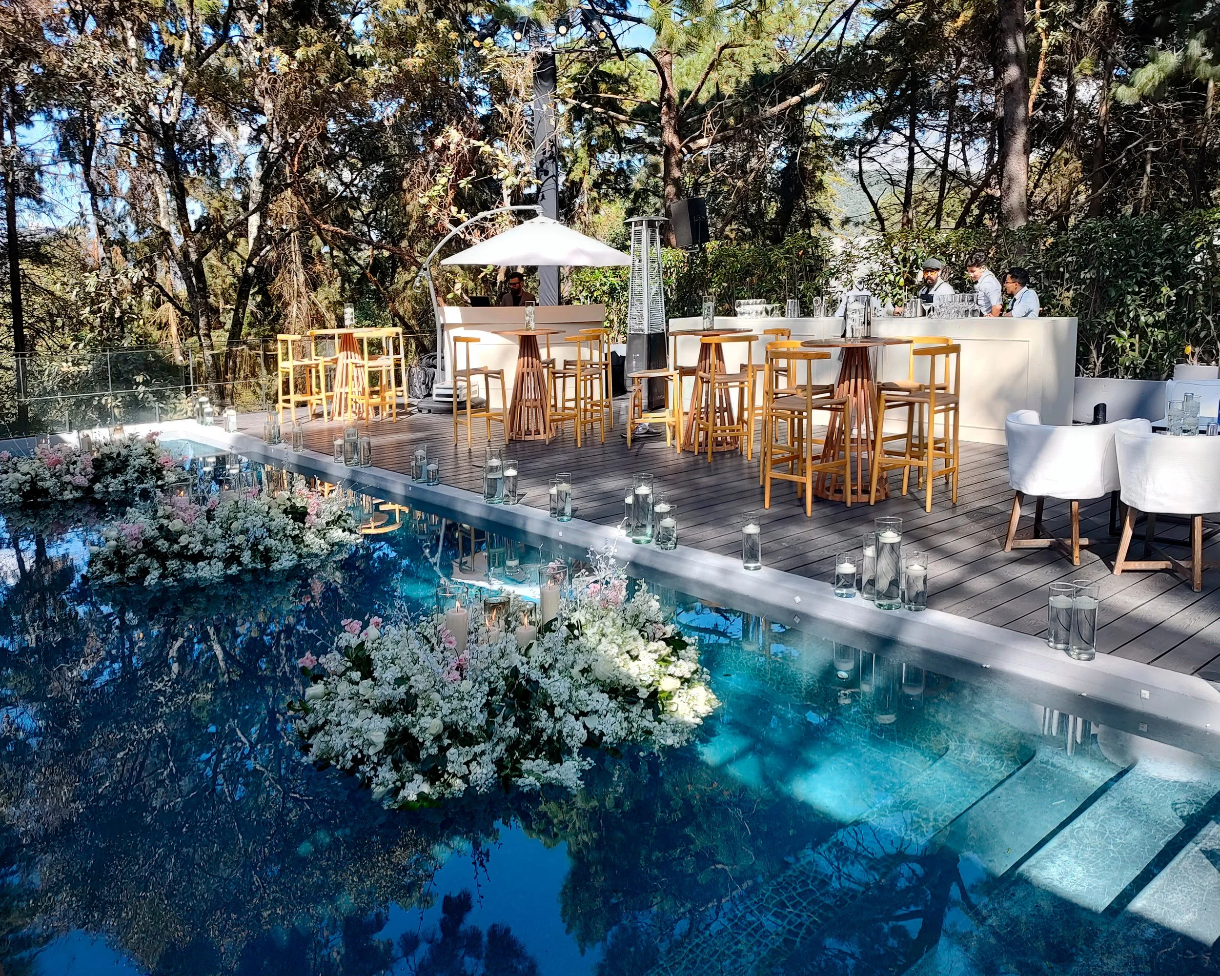Decoración de evento en una terraza cerca de una piscina con flores blancas, mesas y sillas de madera y blanco, y un bar con personal sirviendo bebidas.