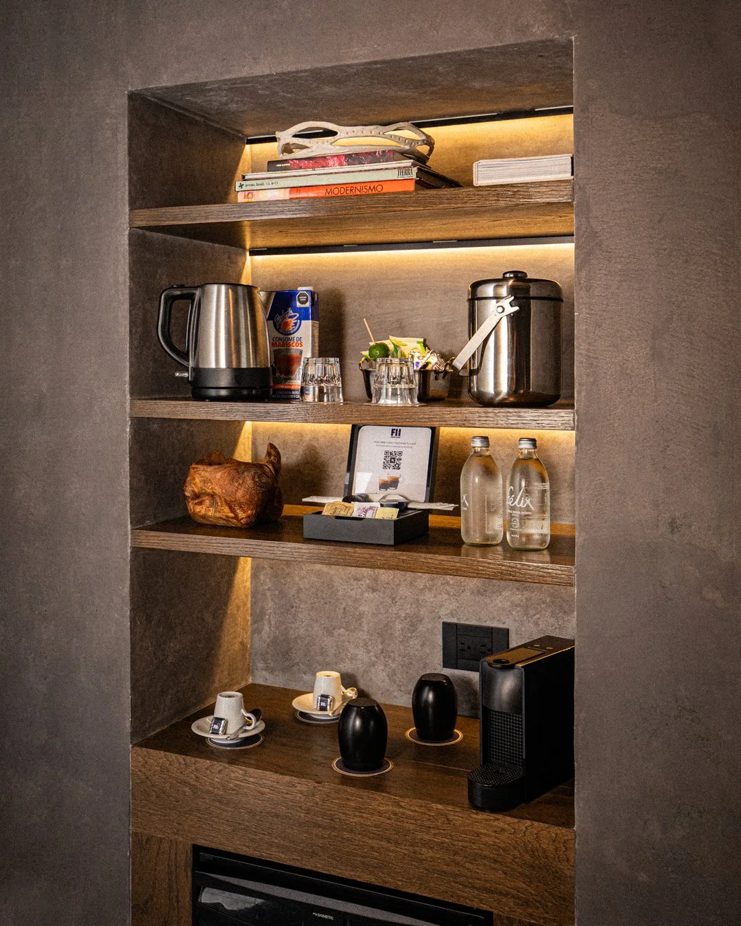 Estantes de madera con utensilios de cocina, tazas, botellas de agua, máquina de café y otros objetos en una pared de cerámica gris.