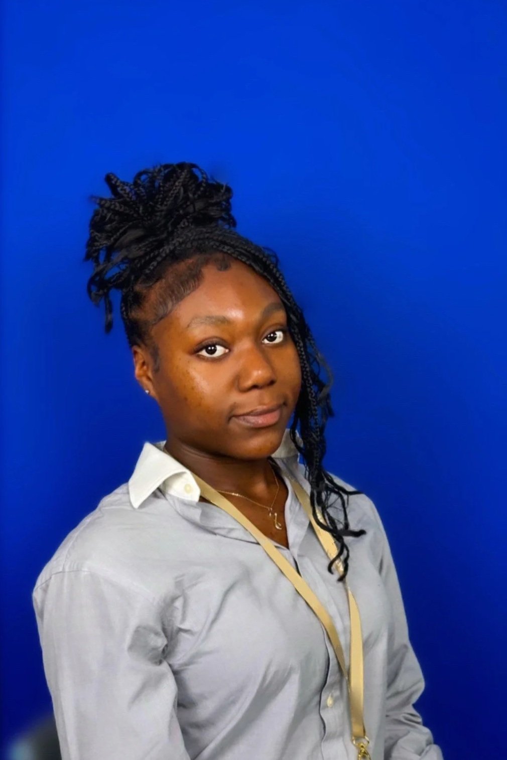 A black woman with braided long hair, wearing a shirt, standing against a bright blue background.
