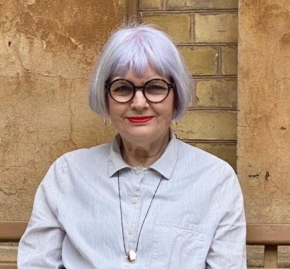 A silver, short haired woman with black rounded glasses, wearing a striped shirt and a necklace.