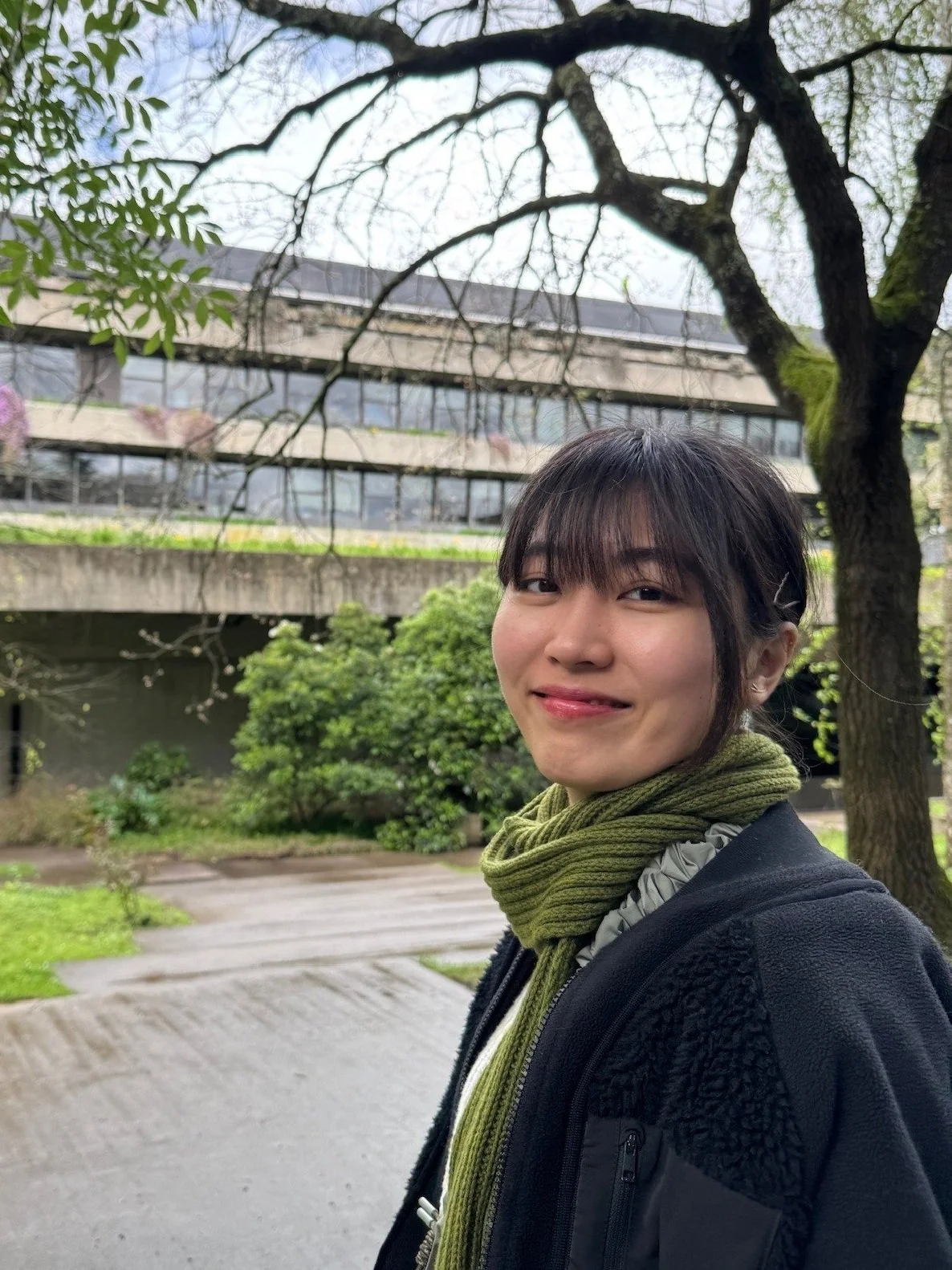 young woman with bangs and hair tied up in a bun. Wearing a collared shirt and grey coat, against a light blue background.