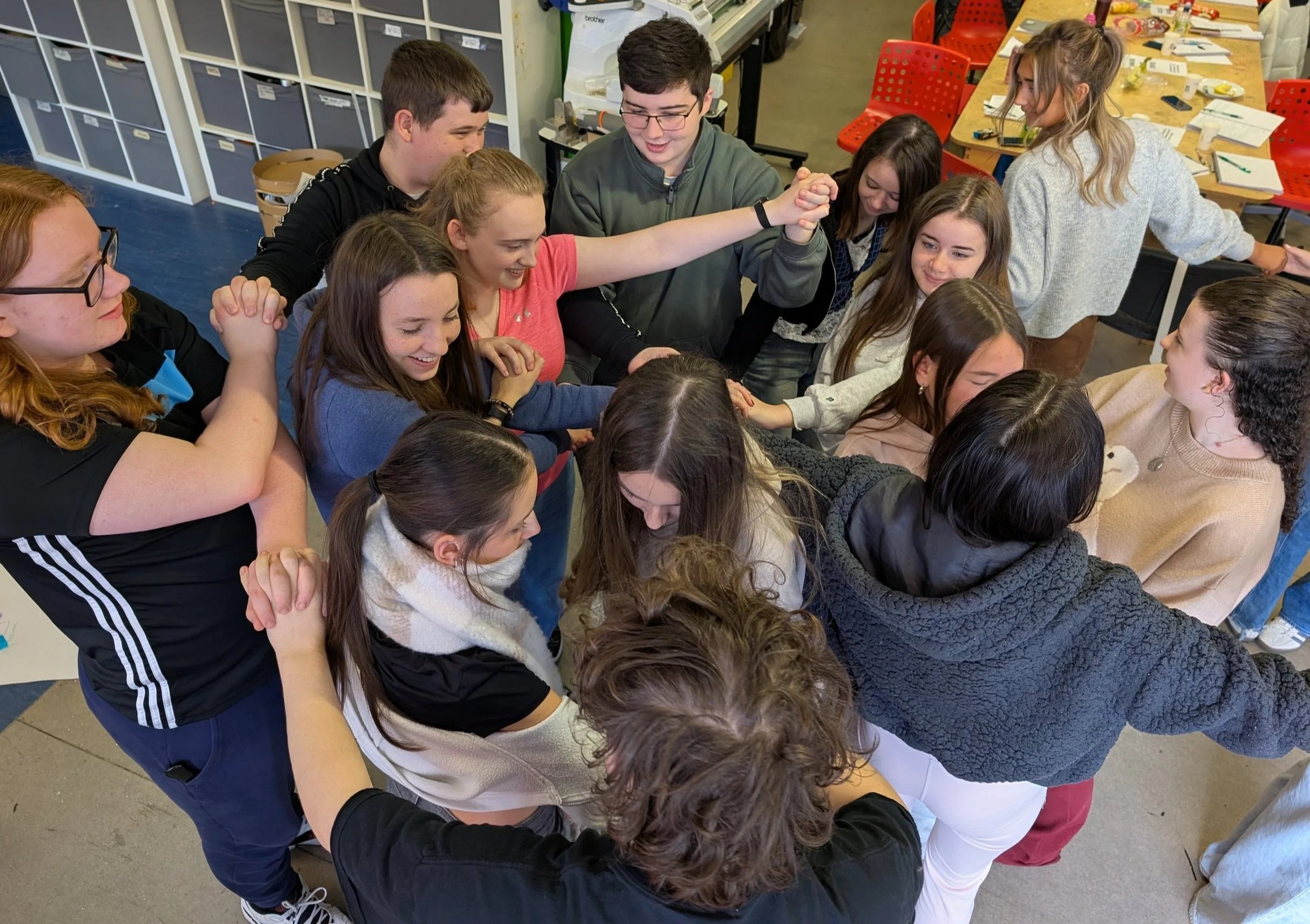 A group of young people playing a game - holding hands with overlapping arms, in a workshop setting.