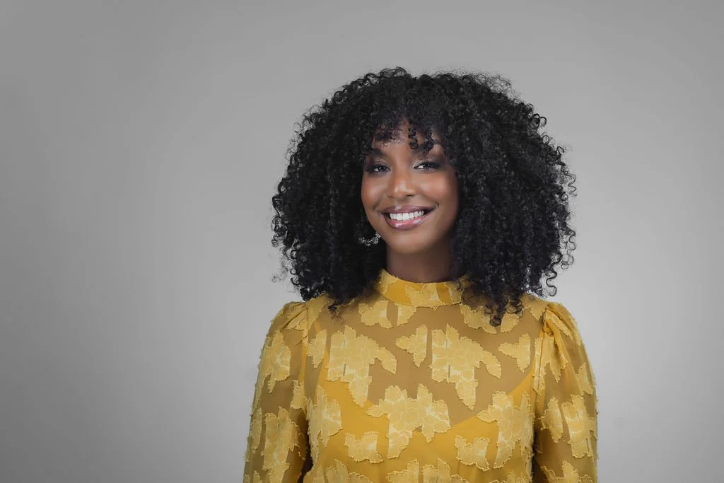 A black woman with curly hair, in a yellow patterned top.