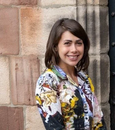 A short haired woman wearing a patterened shirt, standing in front a brick building.