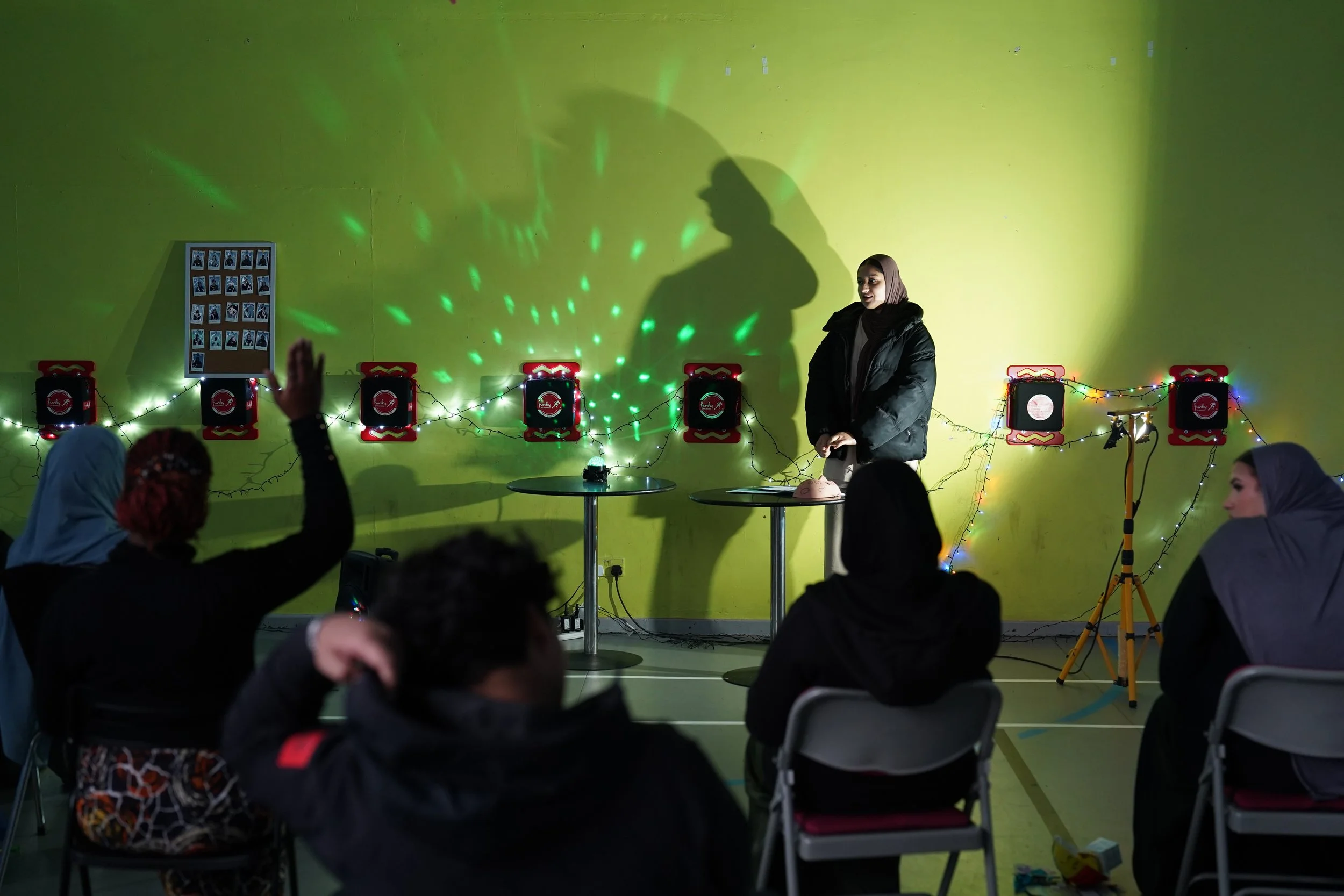 A girl sharing in front of a crowd of young people in a atmospheric room lit by fairylights. One girl raise their hand.