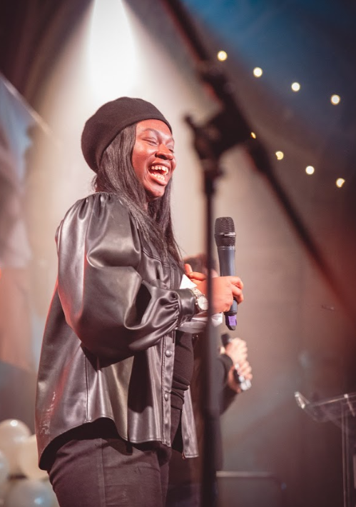 A black woman with a balck beret and a black leather jacket, smiling and holding a mic,