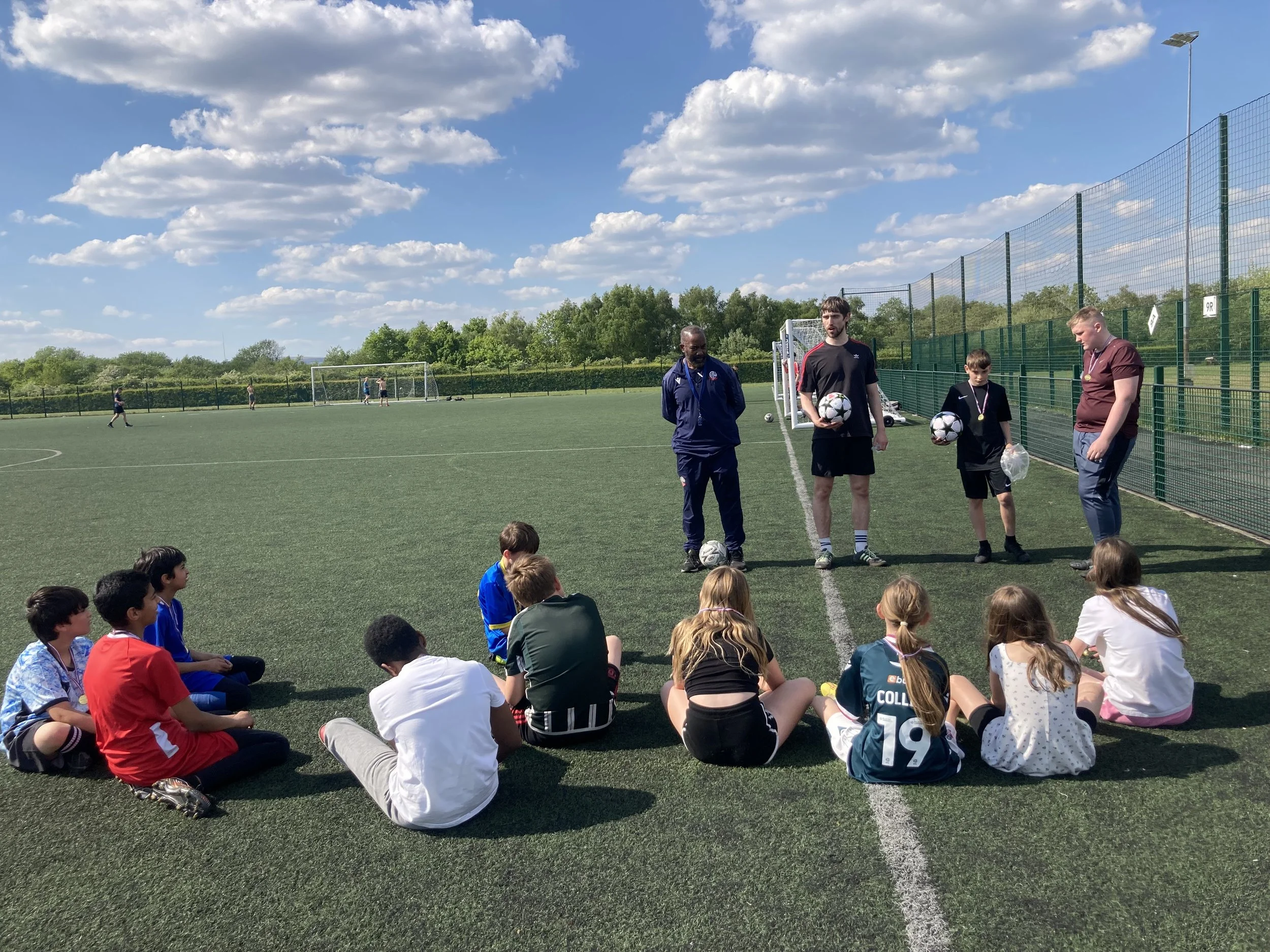 Children sitting on the turf field listening to coaches during a soccer training session on a sunny day.