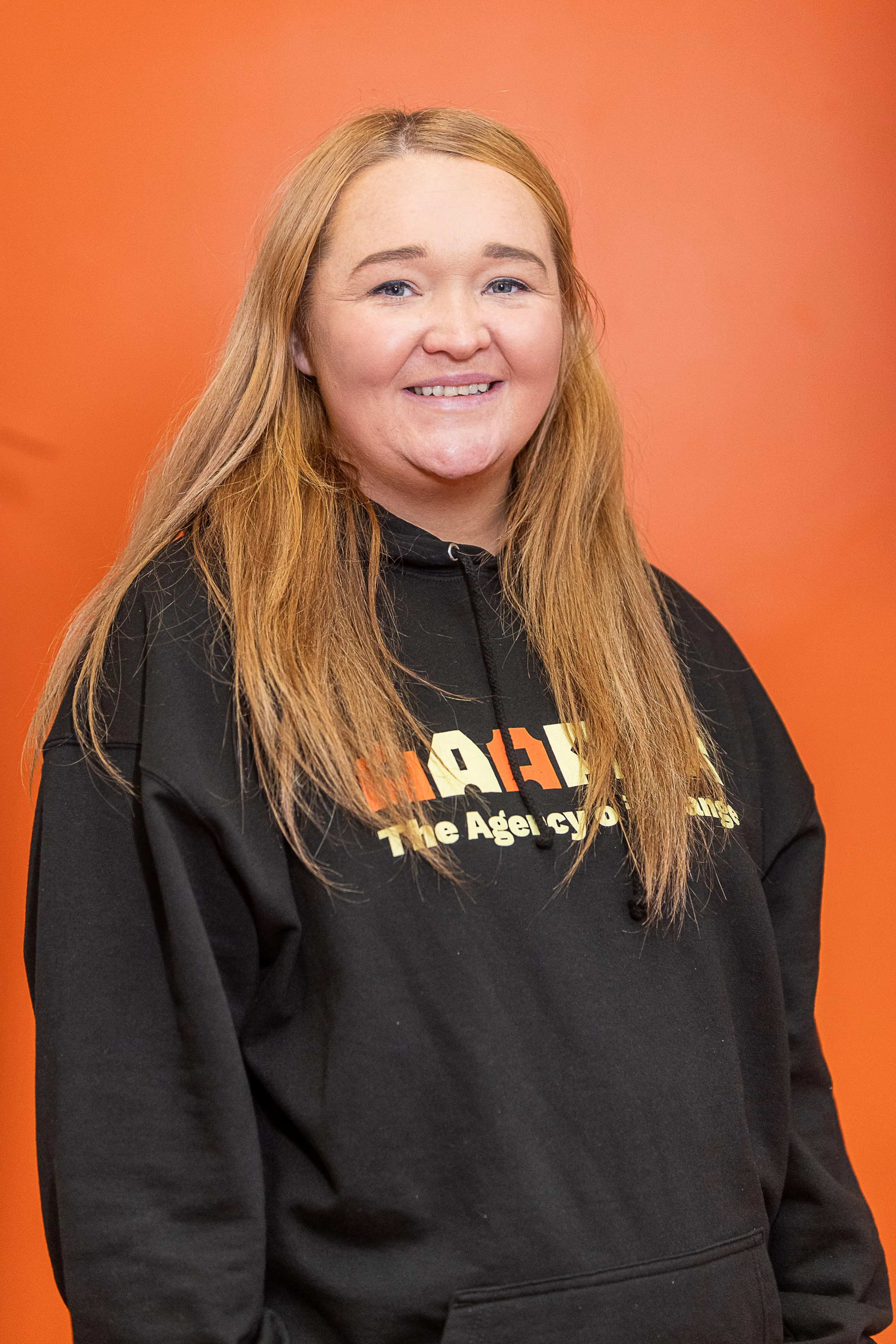 A woman with ginger hair, wearing a hoodie against orange background.