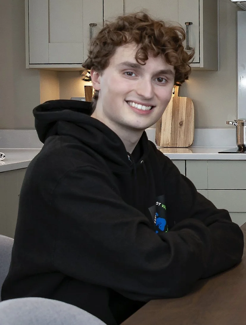 A white young man witth curled hair, wearing a black hoodie. Seated in front of a kitchen.