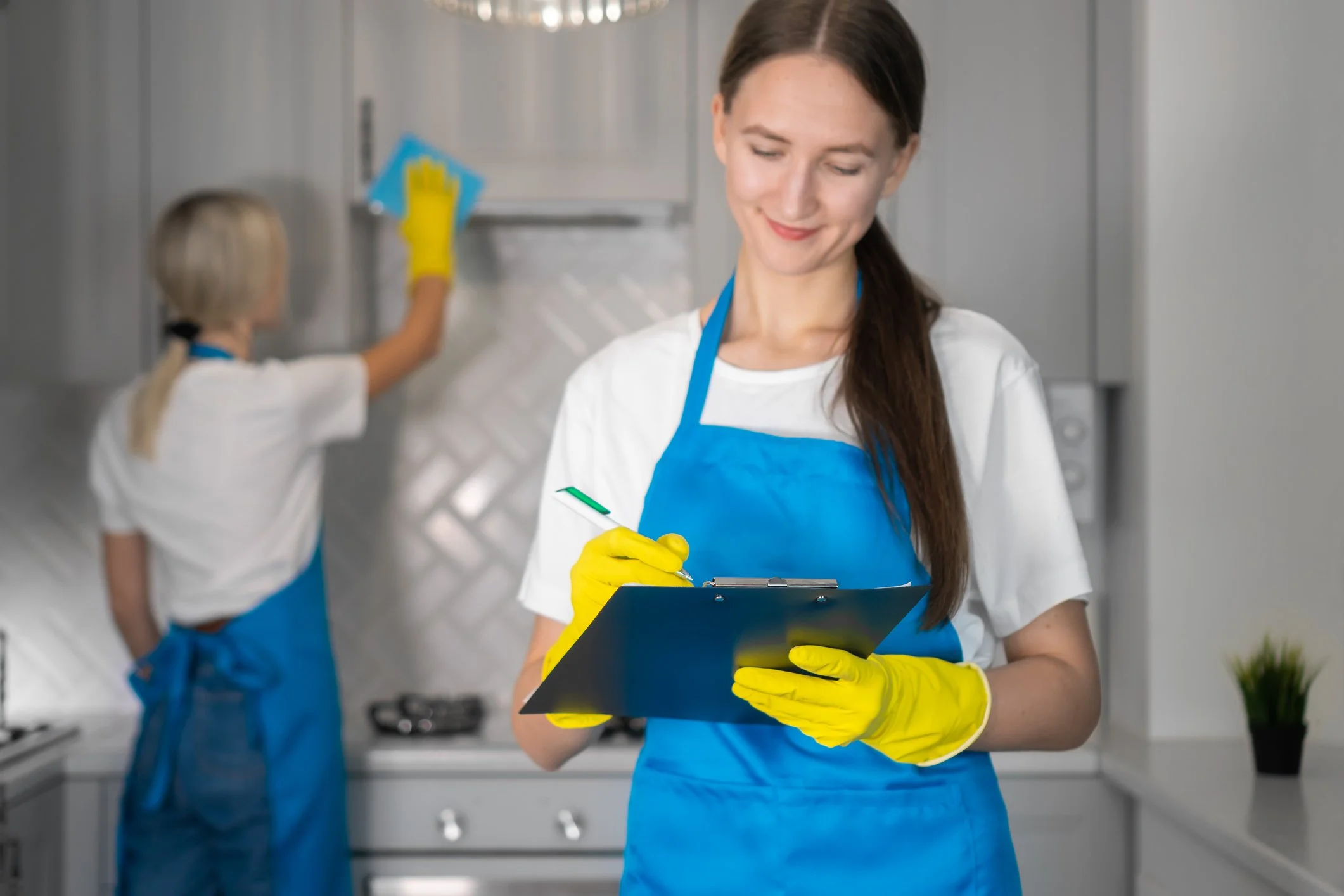 A woman in a white shirt, blue apron, and yellow gloves holding a clipboard and pen, smiling, in a professional kitchen. A young girl in similar attire is cleaning  a commercial Turn Over. for Happyhouse509