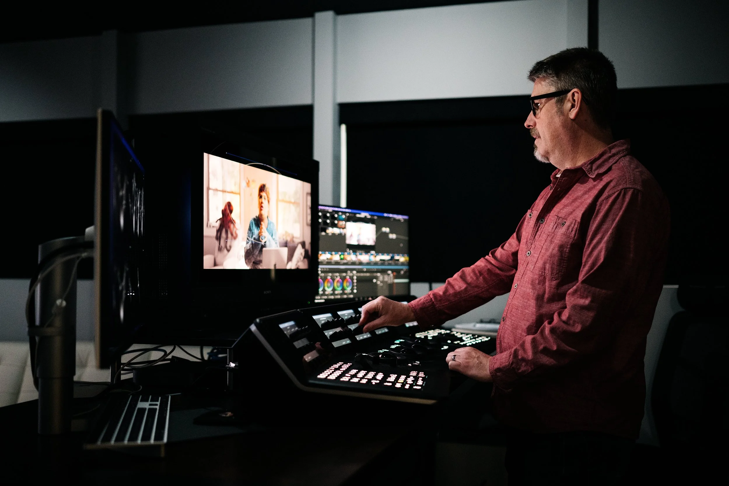 Man operating video editing equipment in a dark editing suite, with multiple monitors displaying video footage and color grading tools.