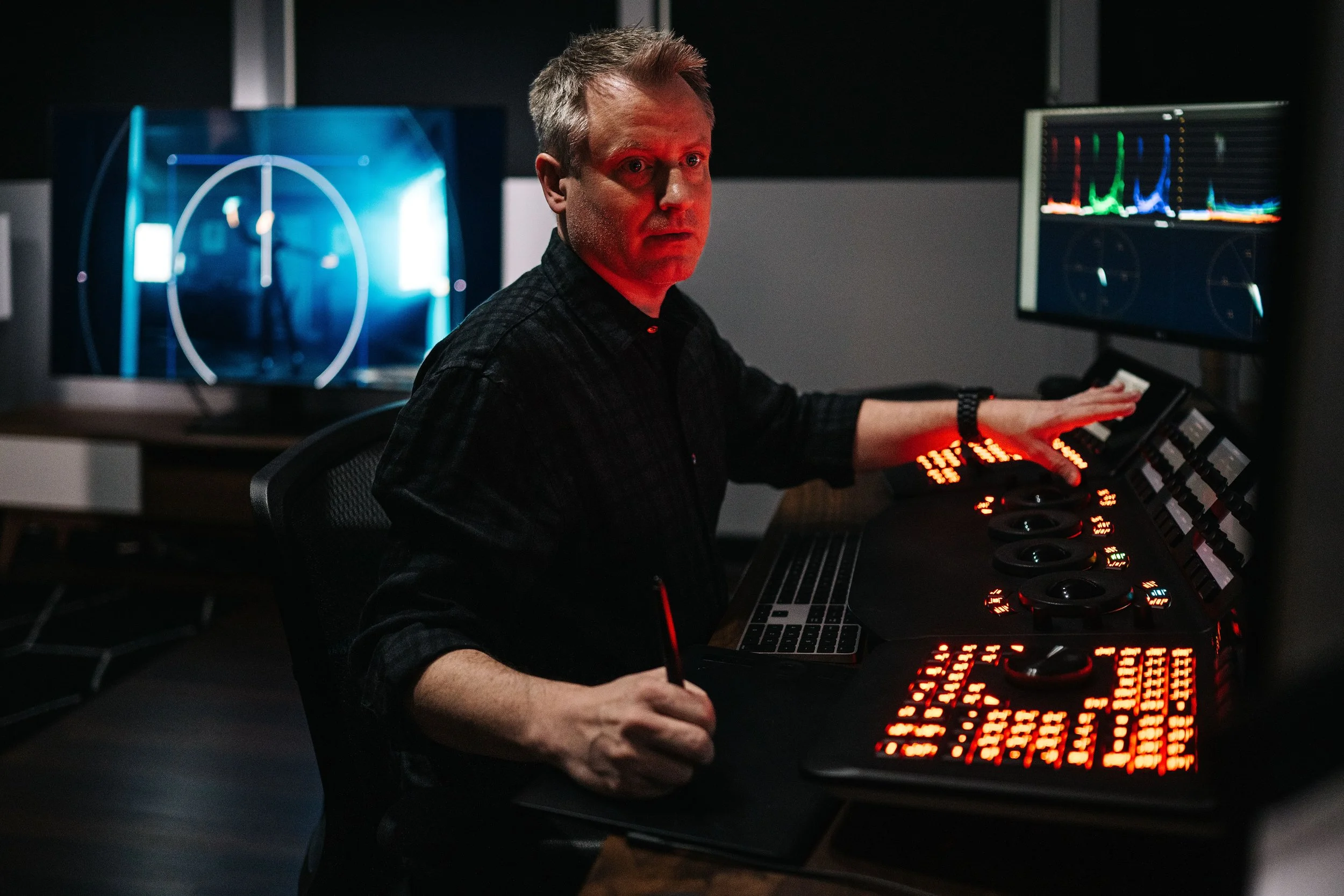 A man sitting at an audio control desk in a dark room, working with multiple monitors displaying waveforms and radar, likely in a professional audio or broadcast environment.