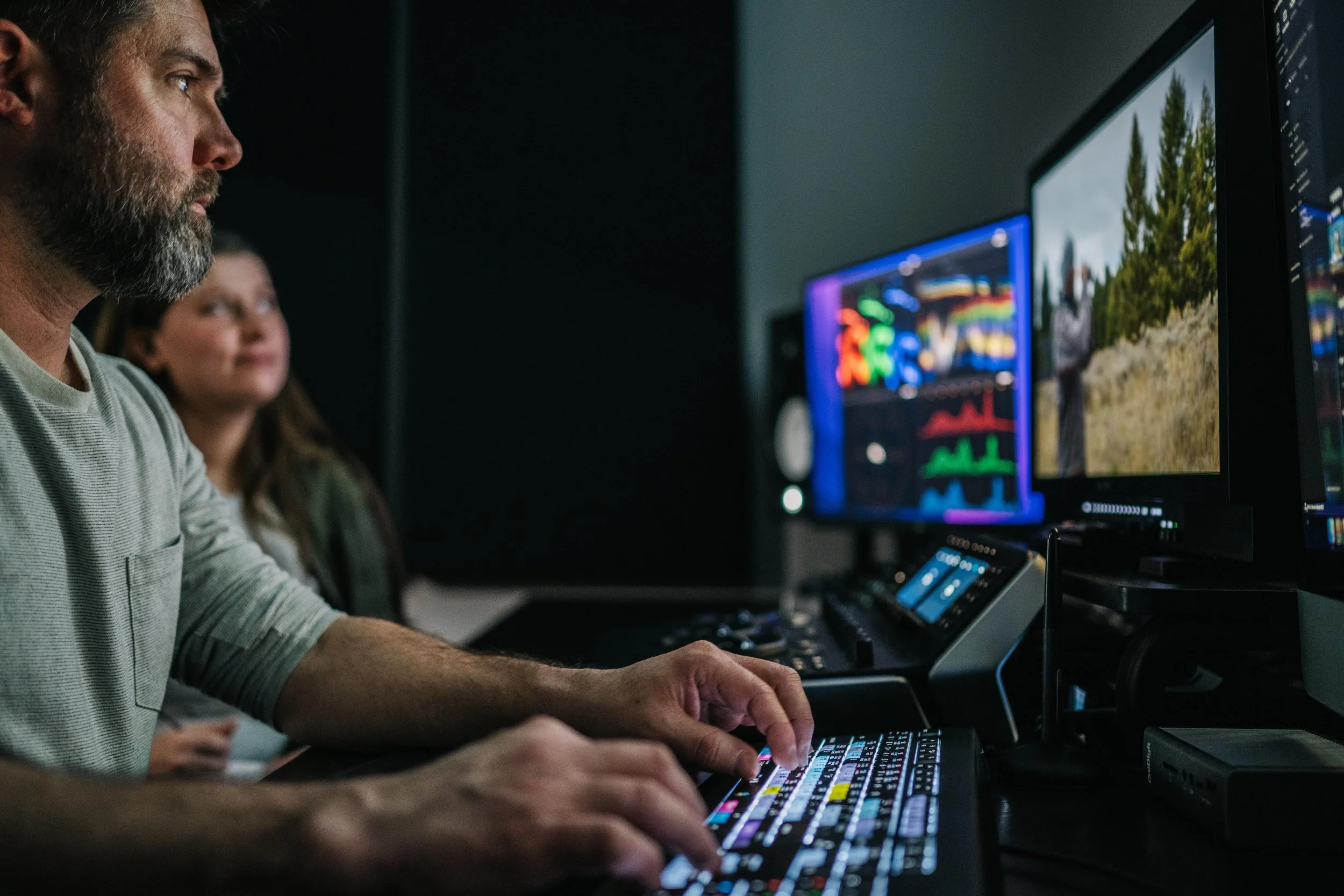 Two people working at a video editing workstation with multiple monitors displaying colorful video editing software and footage.
