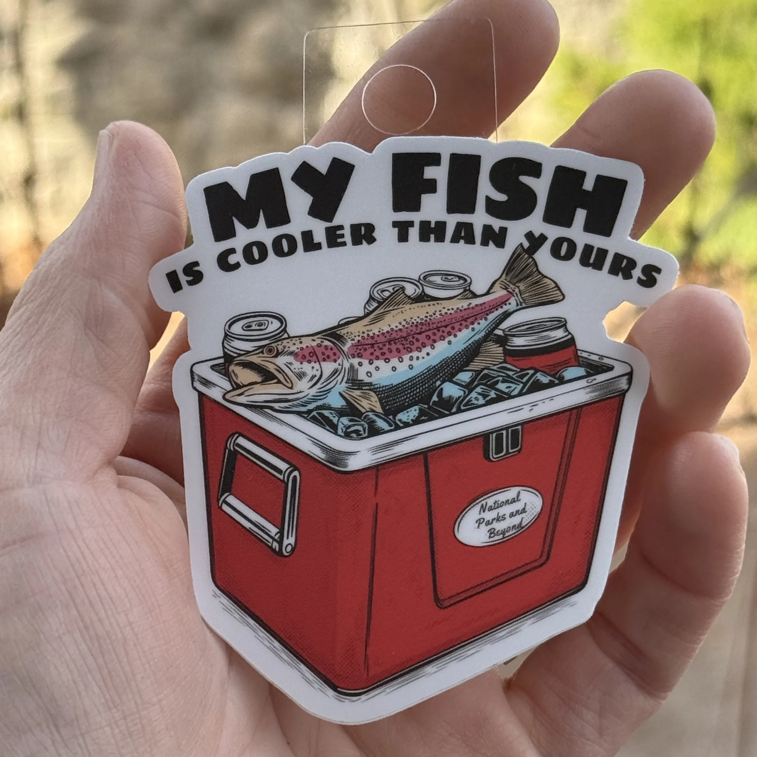 2 inch fishing humor sticker. Featuring a hang tag for display  featuring a red hard sided cooler with ice, cans, and a fish on top with the text “My Fish Is Cooler Than Yours”
