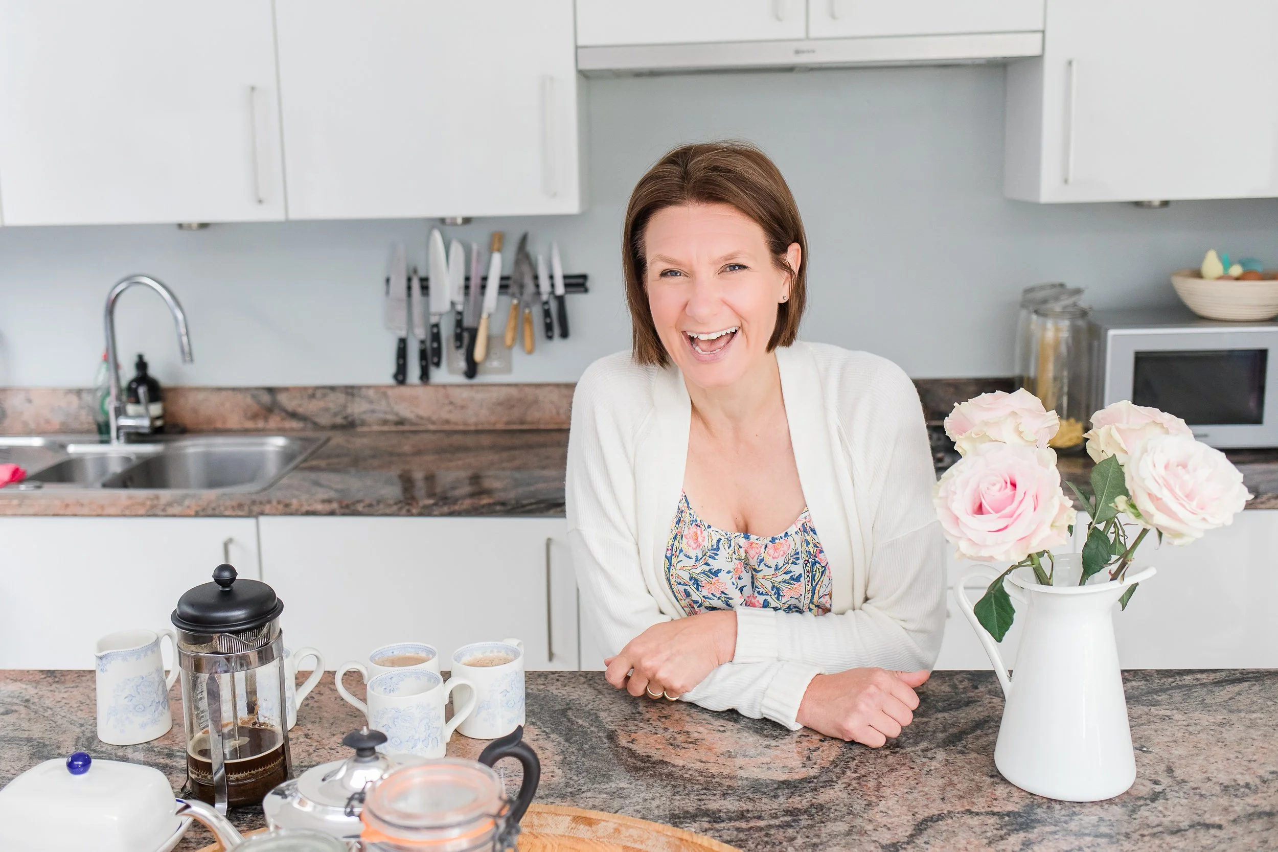 Melissa gauge stands in a kitchen in a floral jumpsuit and white cardigan. Coffee pot and cups are on the work surface and she is standing next to a white jug with pale pink roses in.