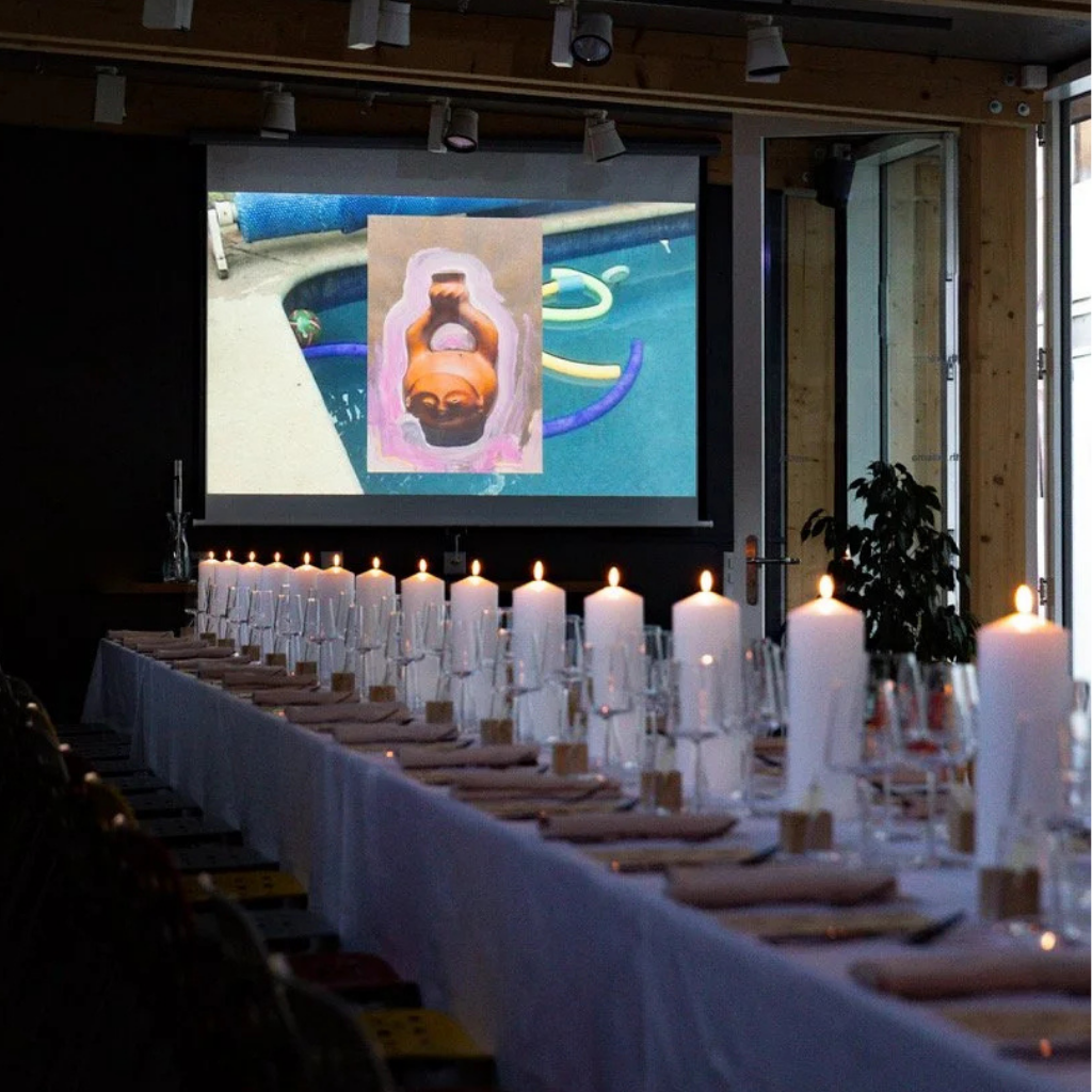 A long banquet table set with candles, glasses, and napkins faces a large projection screen showing a swimming pool and a stretched canvas painting of a woman’s face upside down.
