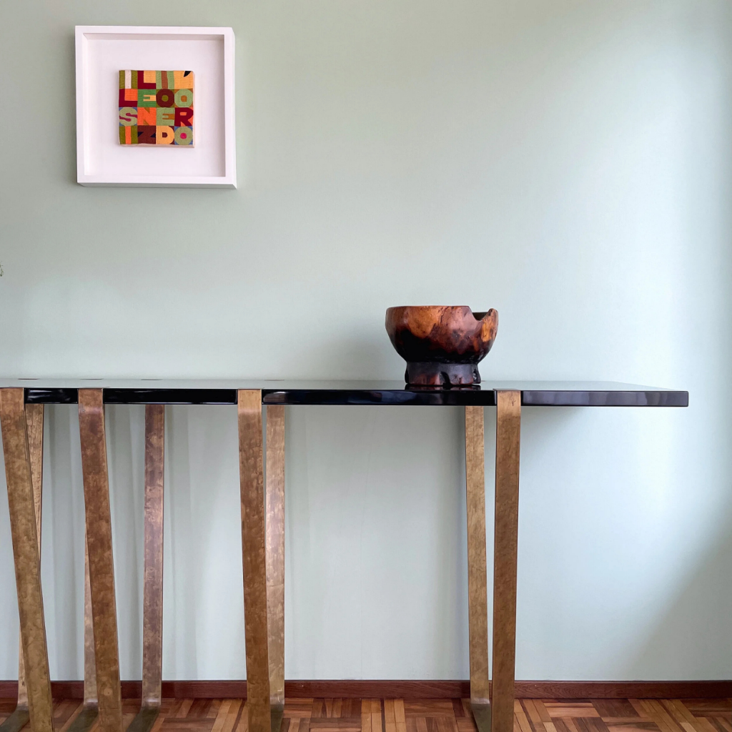 A modern black table with gold legs against a pale green wall. On the table, there's a wooden bowl. Above, a framed colorful textile art piece hangs on the wall.