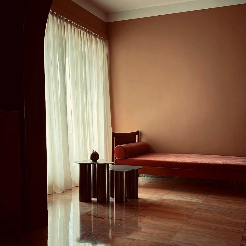 A minimalist interior with a coral-colored daybed, a small round side table with a decorative item, and sheer curtains allowing natural light into the room.
