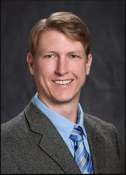 Portrait of a smiling young man in a gray suit jacket and blue shirt with a blue striped tie against a dark background.