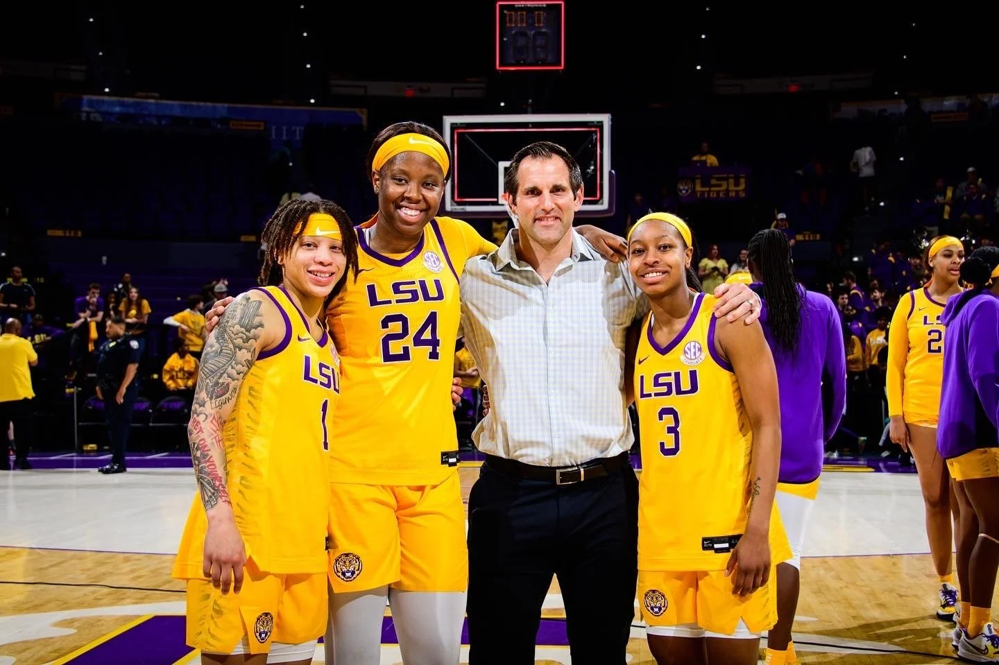 Four women basketball players in yellow LSU uniforms and one man pose on a basketball court, smiling for the camera, after a game.