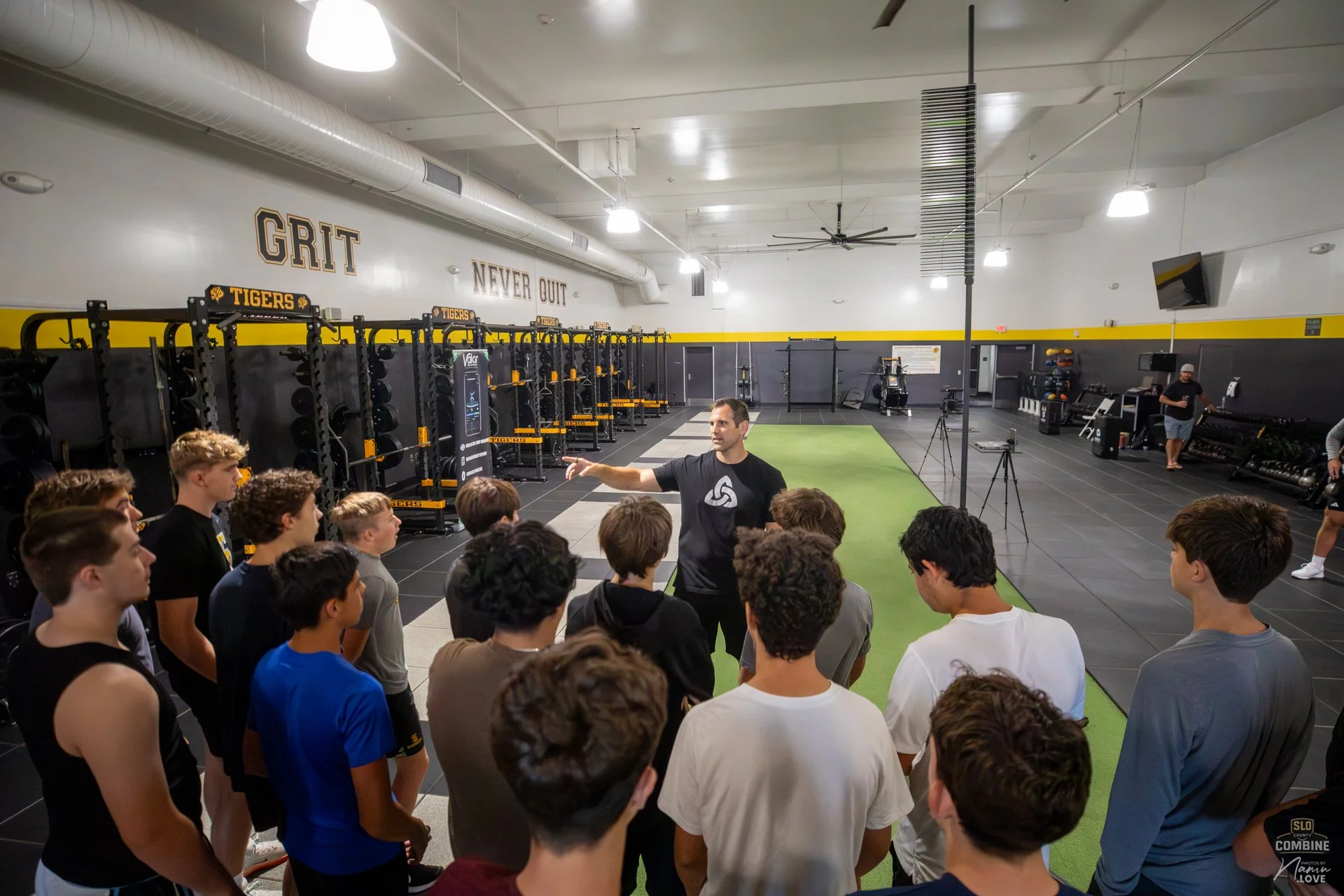 A group of young men attentively listening to a coach inside a gym with black and yellow walls, weight racks, and exercise equipment, as the coach gestures with his right hand during a discussion or training session.