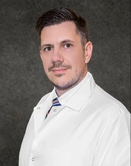Portrait of a man in a white coat with a striped tie against a dark background.