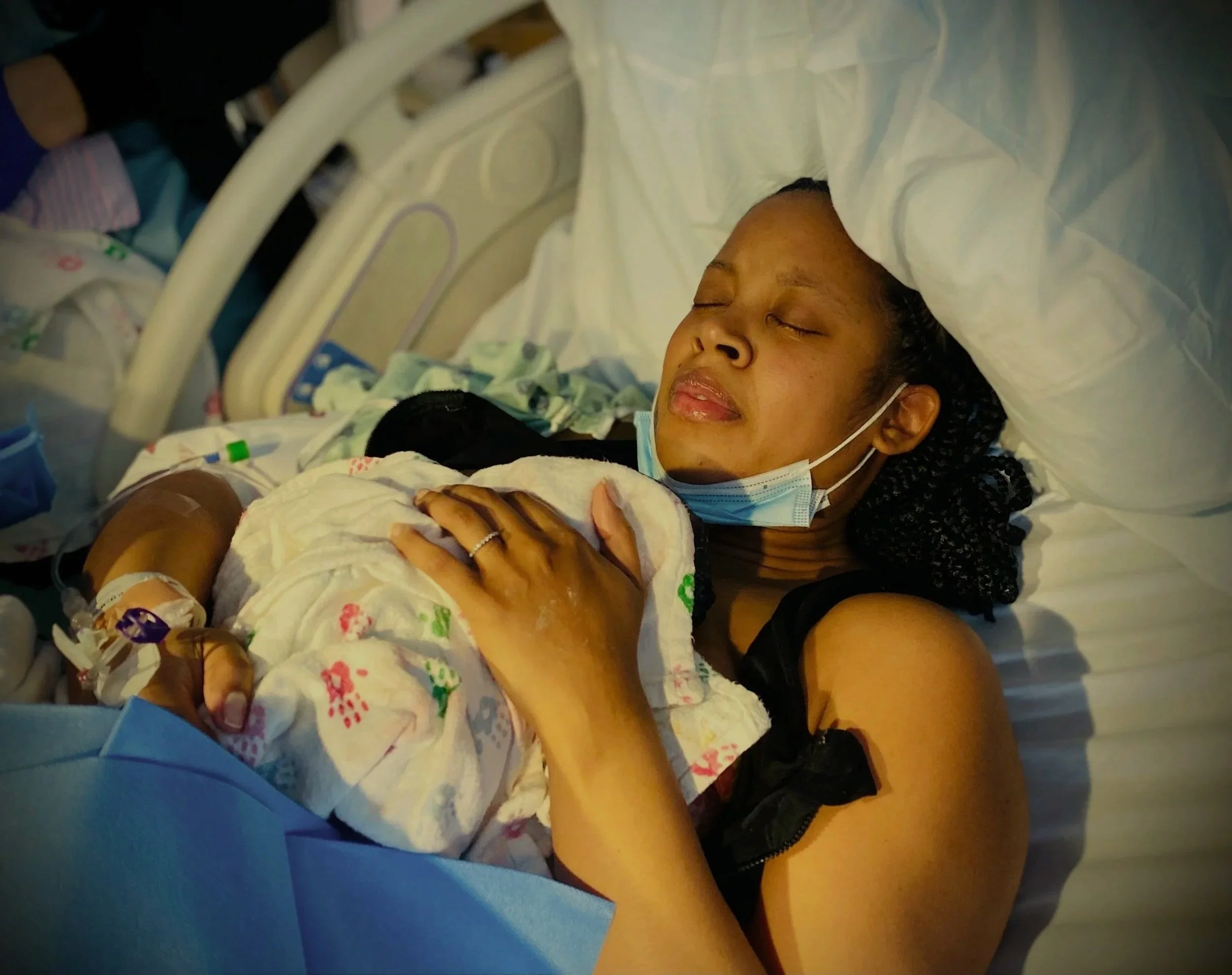 Woman lying in a hospital bed with closed eyes, wearing a face mask and holding a newborn baby wrapped in a hospital blanket, hospital equipment visible in the background.