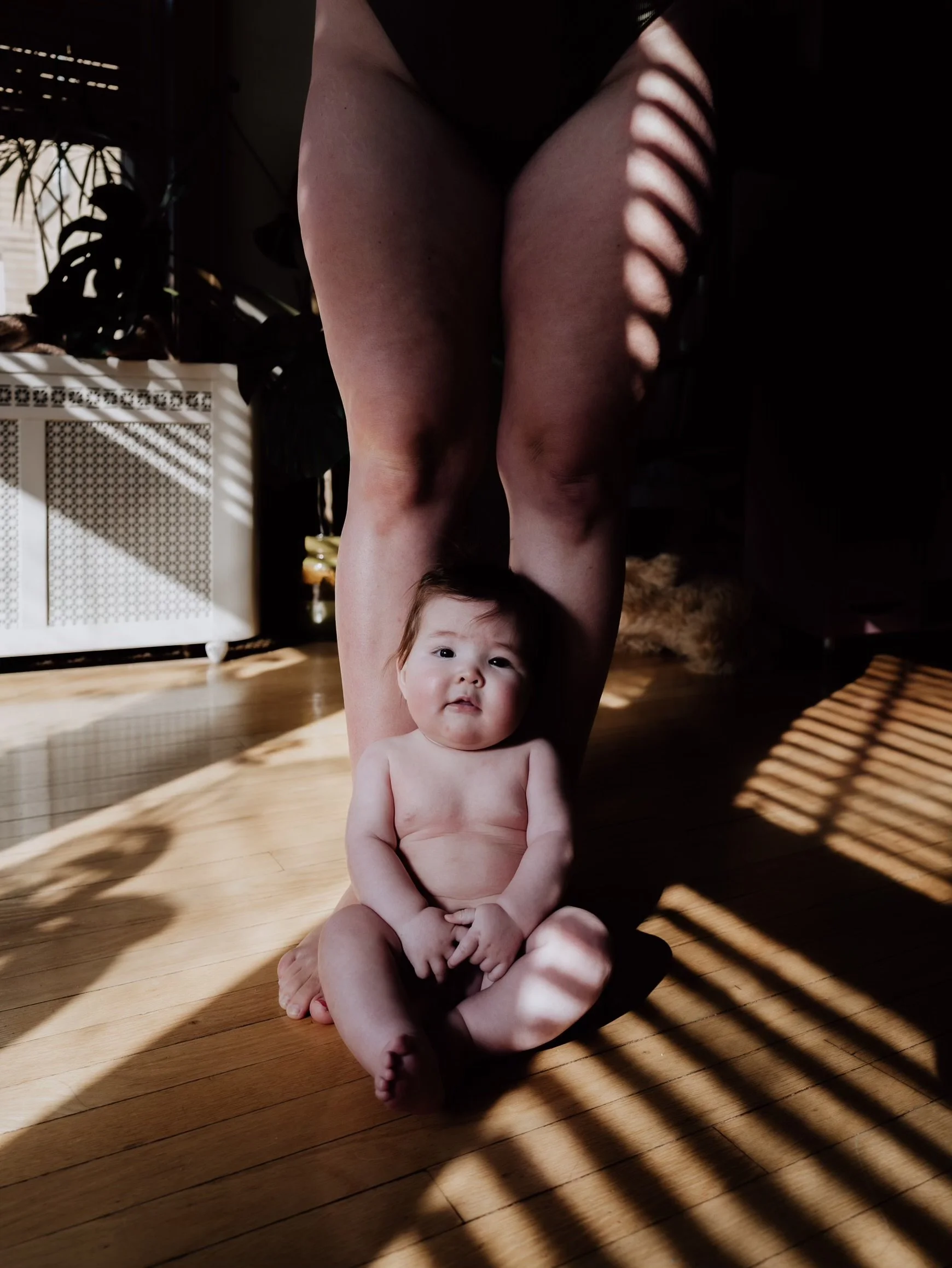 A baby sitting on a wooden floor, with sunlight and shadow patterns through window blinds casting across their body, and their hair and facial expression visible.