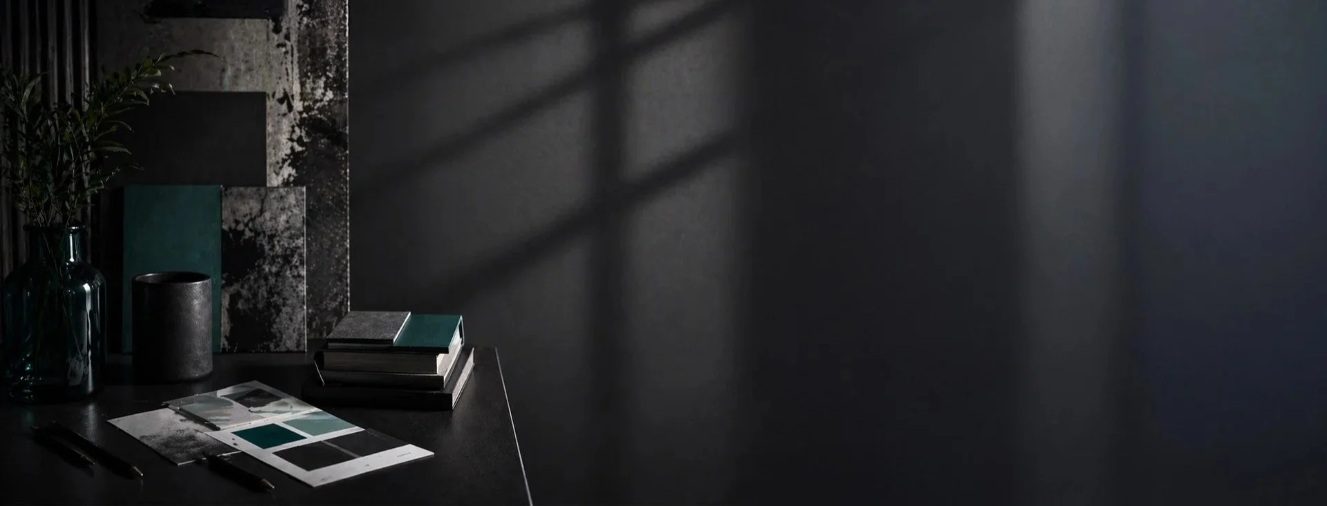 A dark, modern workspace with a black desk, a green plant in a glass vase, stacked color sample books, and paint swatches, with shadows cast on a dark wall.