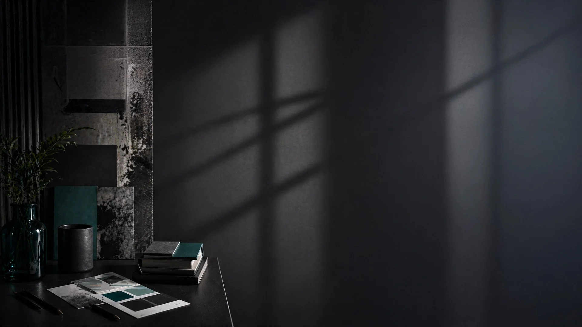 A dark room with a black table displaying color swatches, books, and a pen, with a gray textured wall and shadows cast by window blinds.