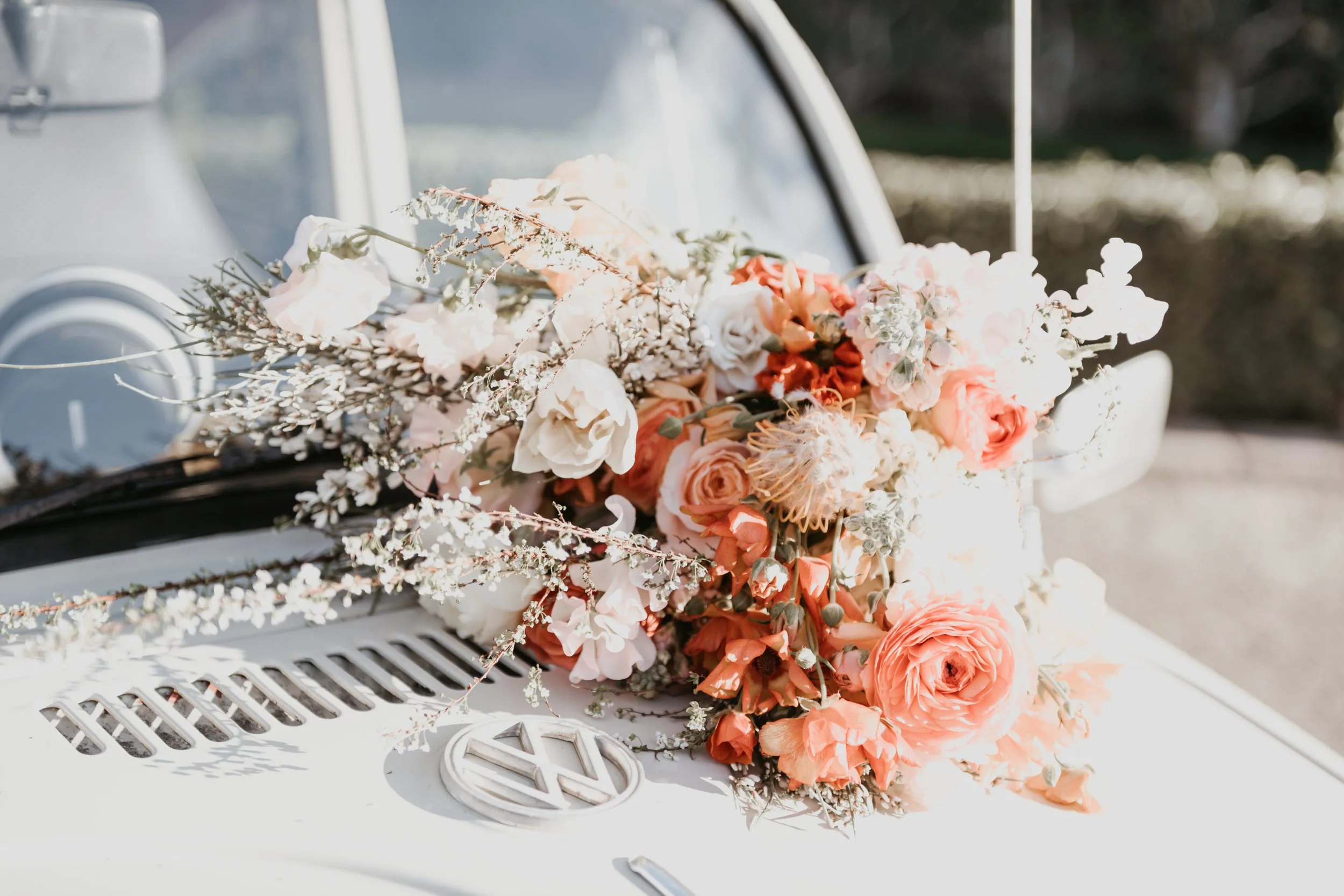 A vintage white car decorated with a large, lush bouquet of light pink, peach, and white flowers, along with greenery and small white blossoms, on the hood for a wedding or special event.