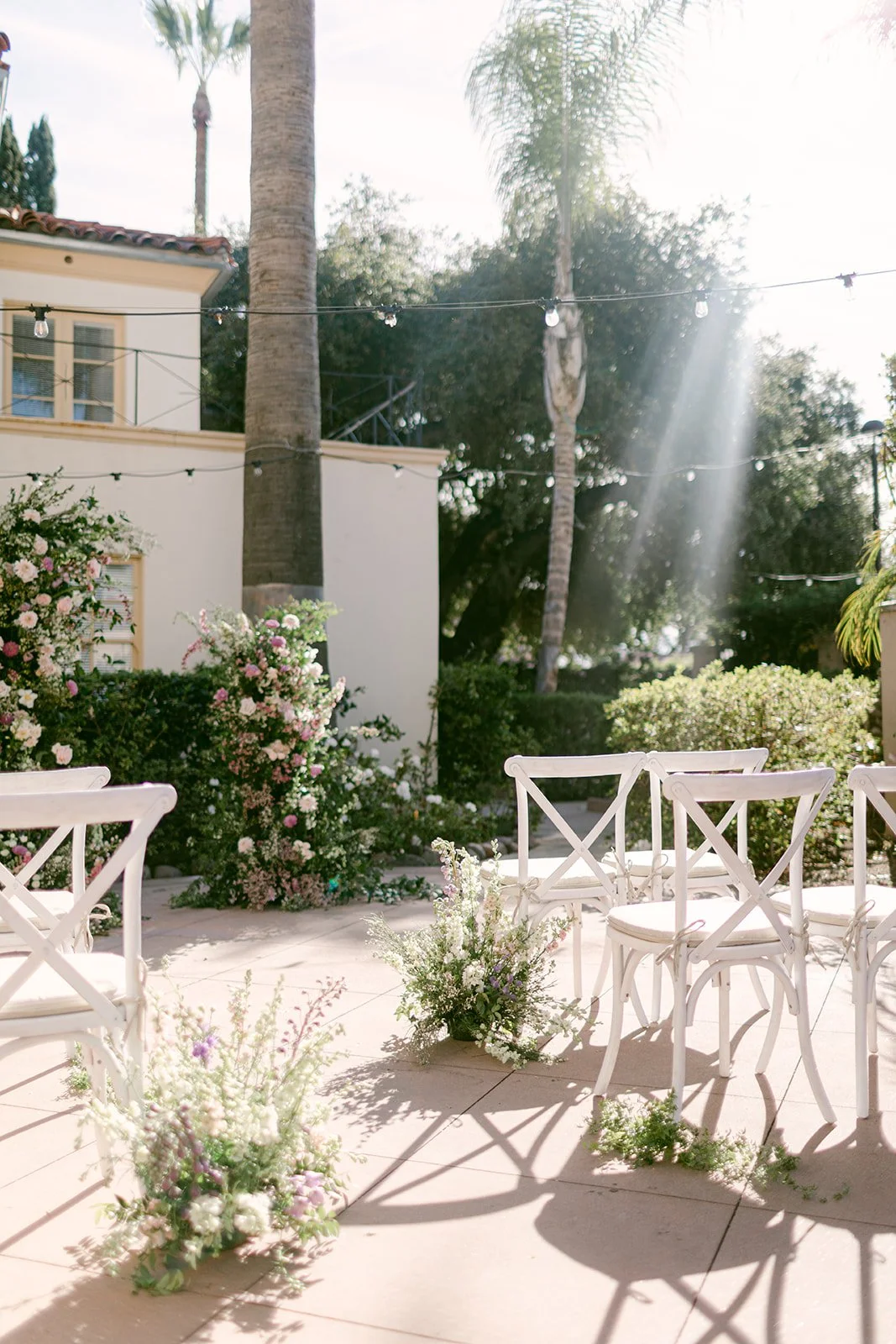 Outdoor patio decorated with white chairs and flower arrangements, with sunlight streaming through trees and string lights overhead.