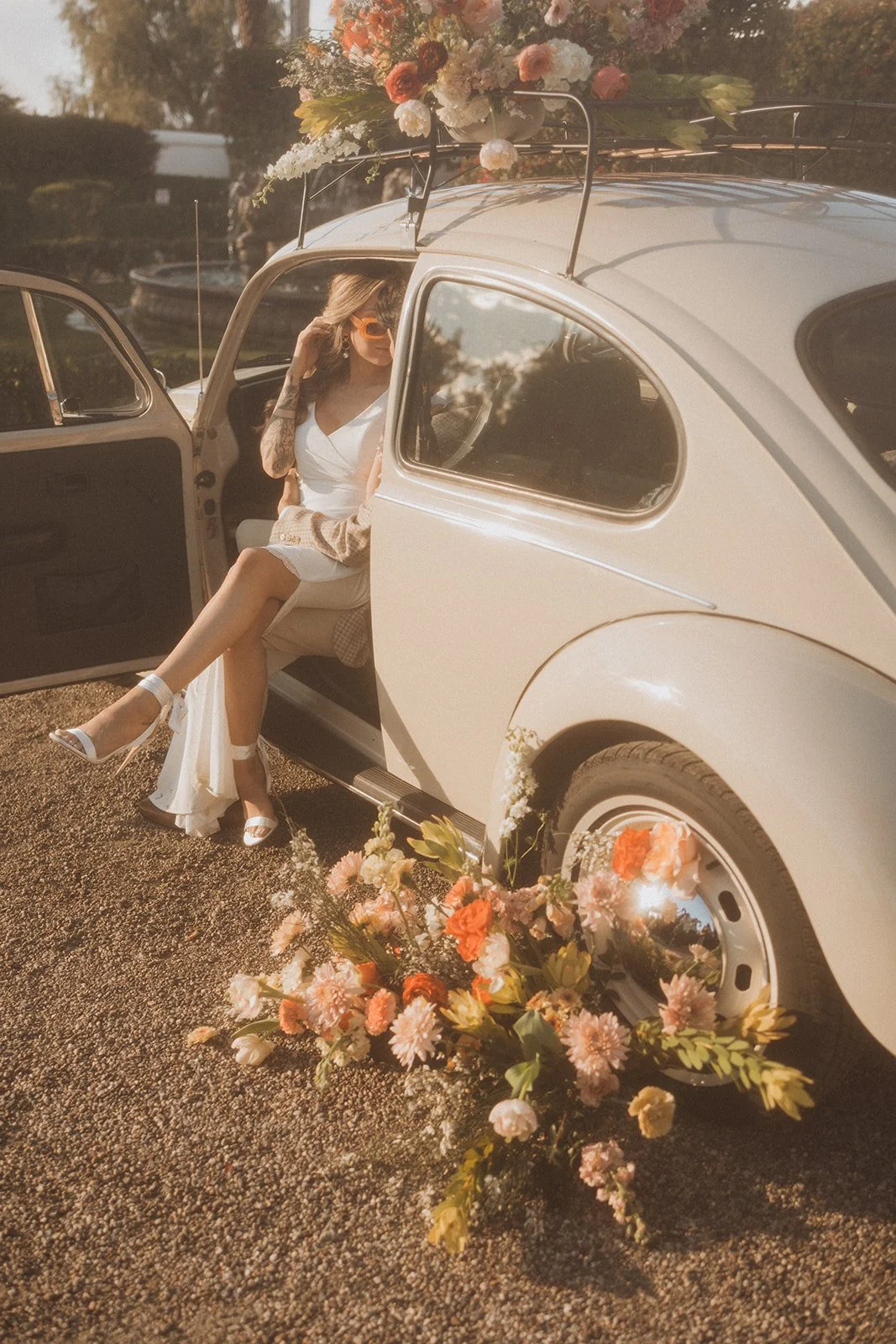 A woman sitting inside a vintage white car decorated with flowers on the sides and on the roof rack, with a bouquet of flowers on the ground nearby, during sunset.