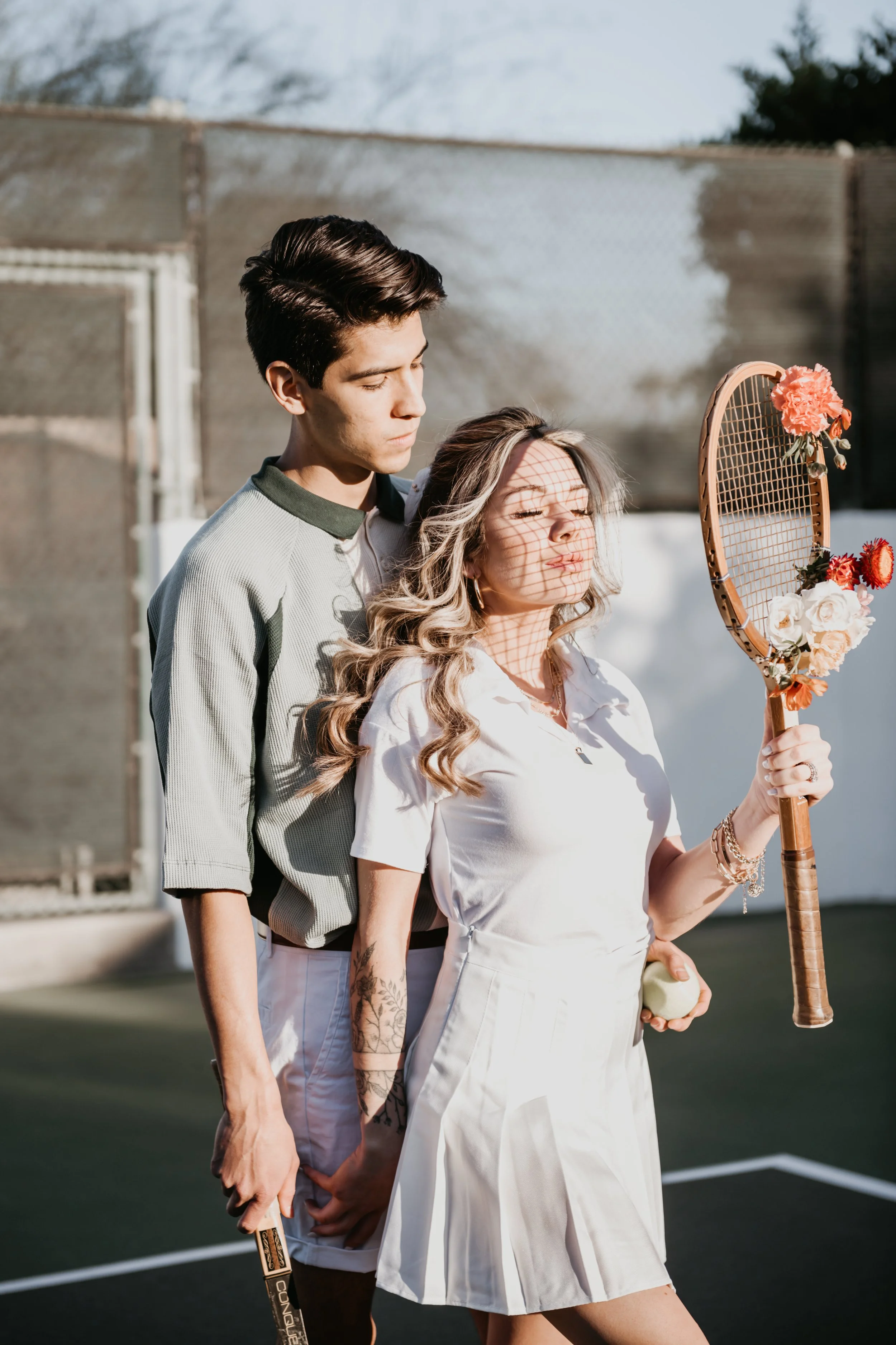 A young man and woman stand close together on an outdoor tennis court, with the woman holding a tennis racket decorated with flowers. The woman has long, wavy hair and tattoos on her arm, and is wearing a white dress. The man has dark, styled hair an