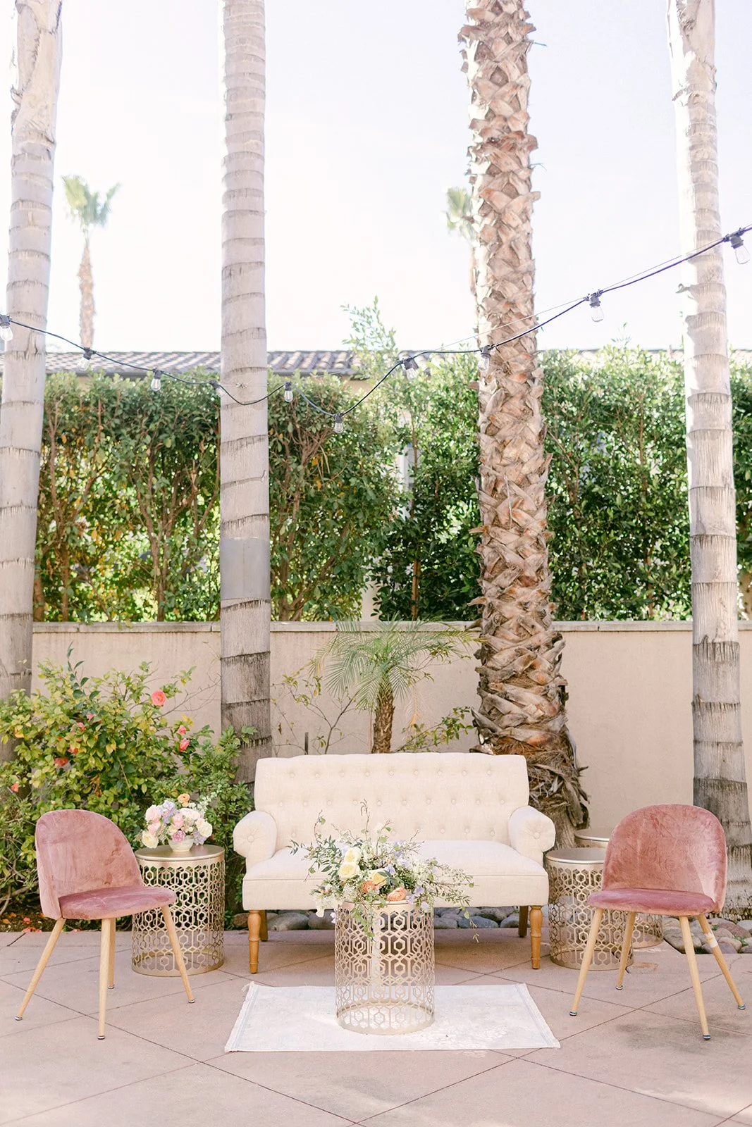 Outdoor patio with pink velvet chairs, a white tufted sofa, floral arrangements, palm trees, and string lights.