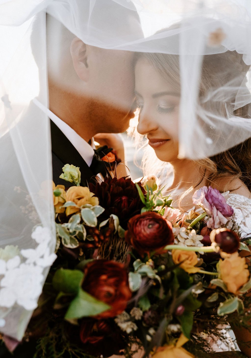 Close-up of a bride and groom smiling with their foreheads touching, wrapped in a delicate veil, holding a colorful bouquet of flowers.