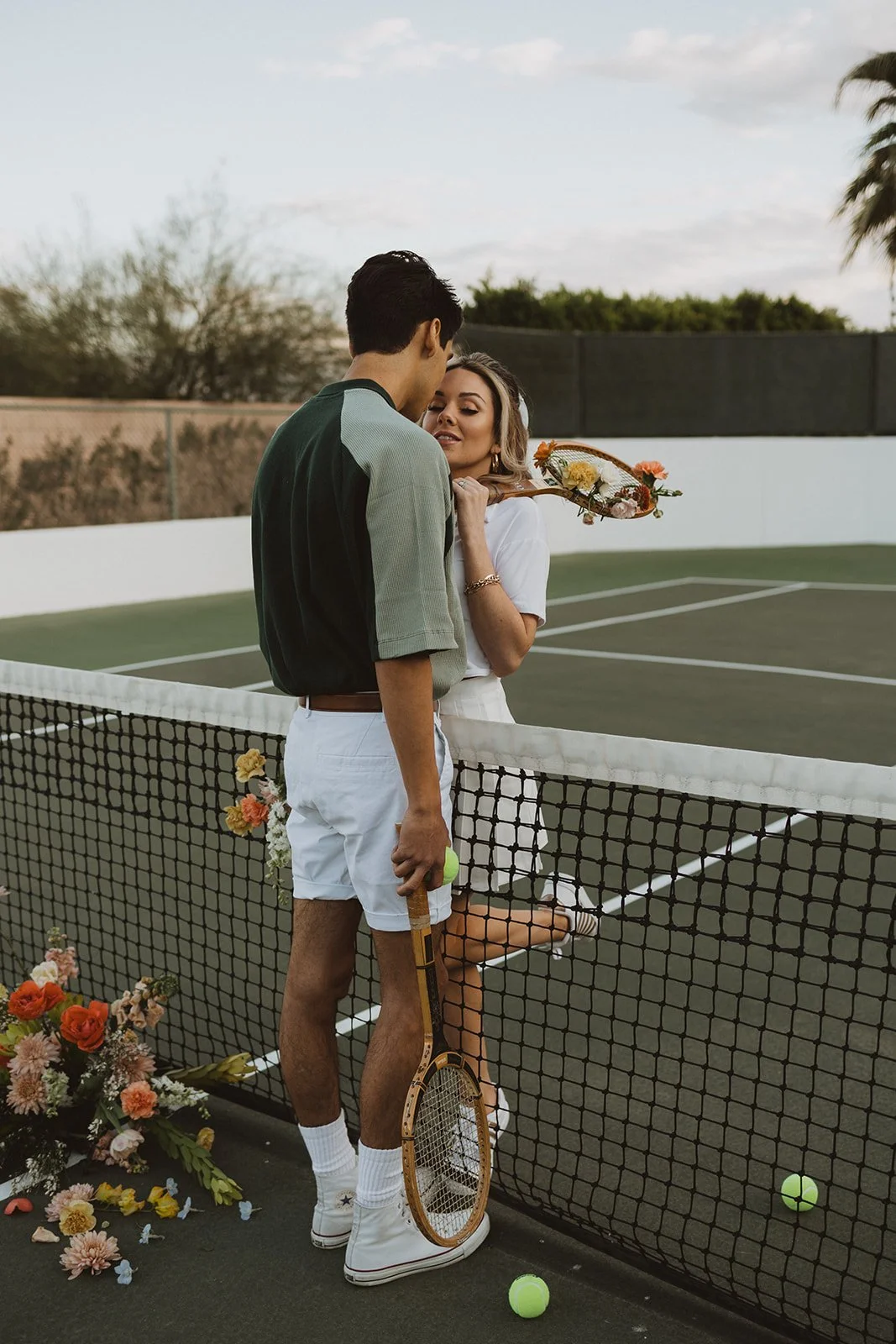 A couple on a tennis court amid a romantic moment, with flowers and tennis balls around, and a woman holding a tennis racket with flowers, sharing an intimate moment.