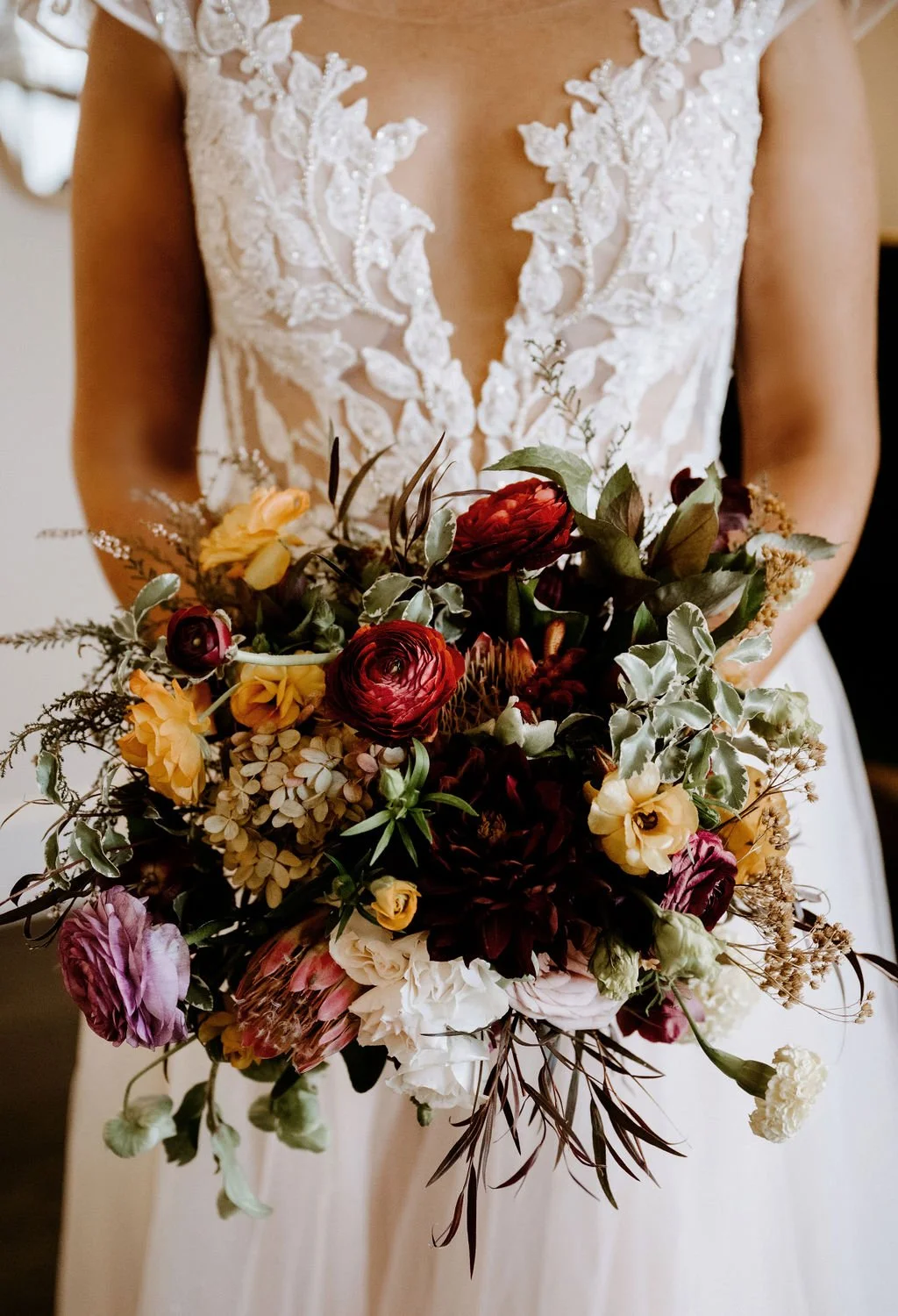 A bride in a white lace wedding gown holding a colorful bouquet of roses, hydrangeas, and greenery.