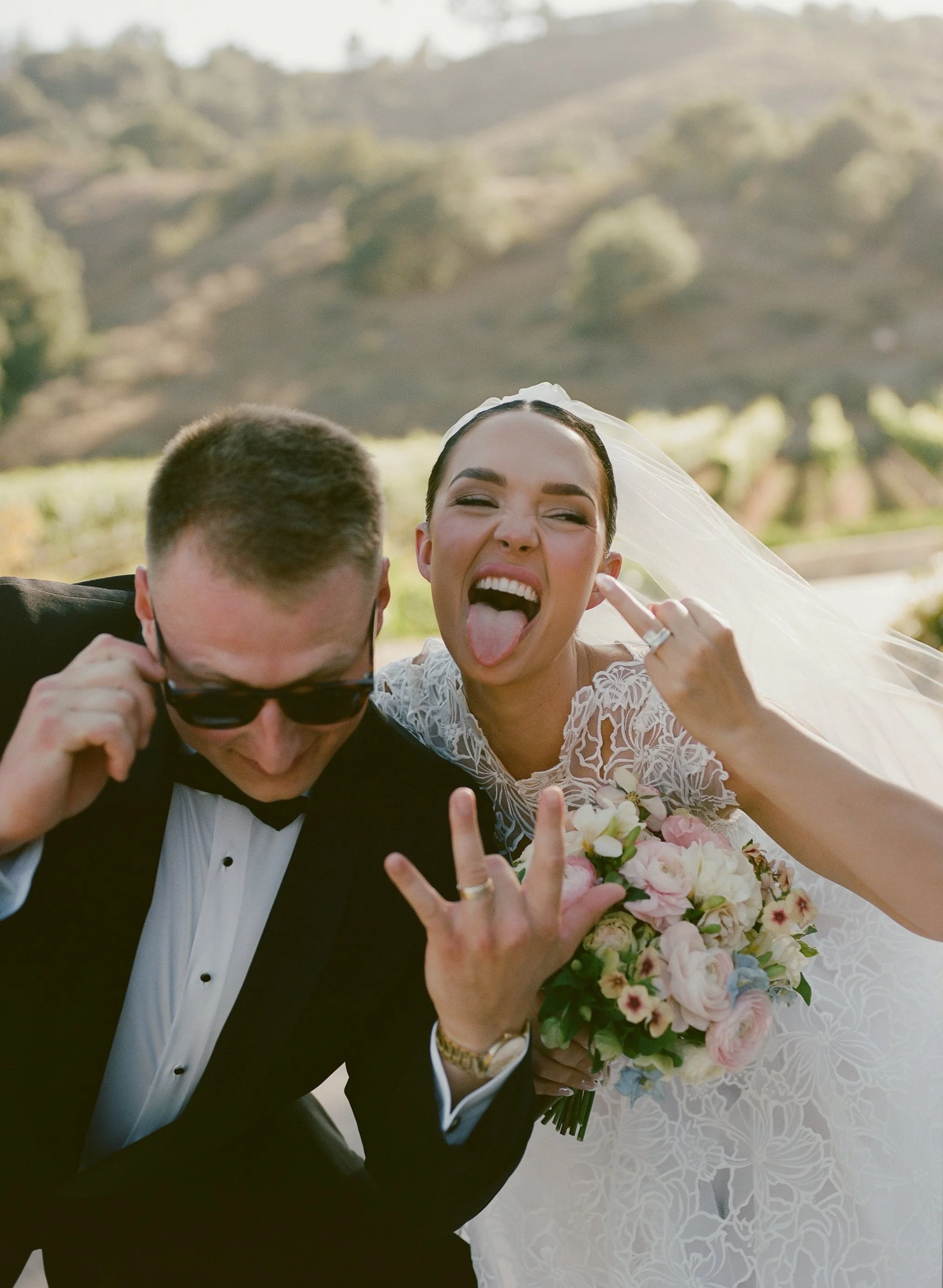 A newlywed couple celebrating outdoors, with the woman sticking out her tongue and the man adjusting his sunglasses. The bride holds a bouquet of pink and white flowers, wearing a white lace wedding dress and veil. The groom wears a black tuxedo with