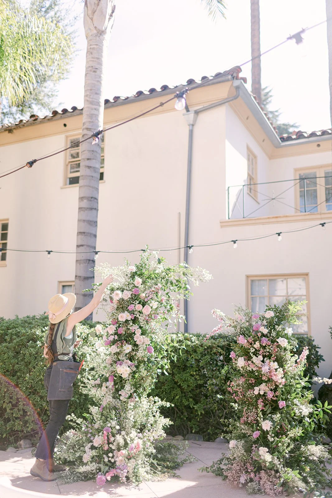 A woman wearing a sun hat and apron arranging pink and white flowers outside near a house with string lights overhead.